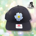 A black Richardson baseball cap with an embroidered white flower with yellow center, displayed against a soft bokeh background with the TundraGeek Creations logo.