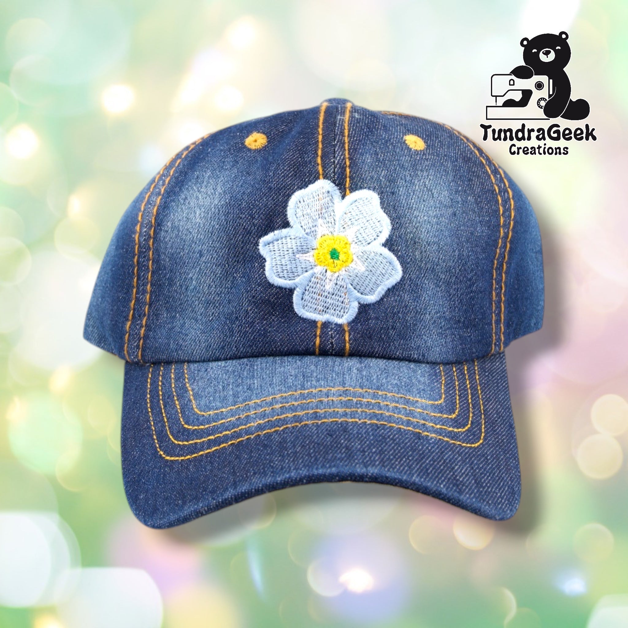A denim baseball cap with gold stitching and a white embroidered daisy flower with yellow center on a soft-focus pastel background.