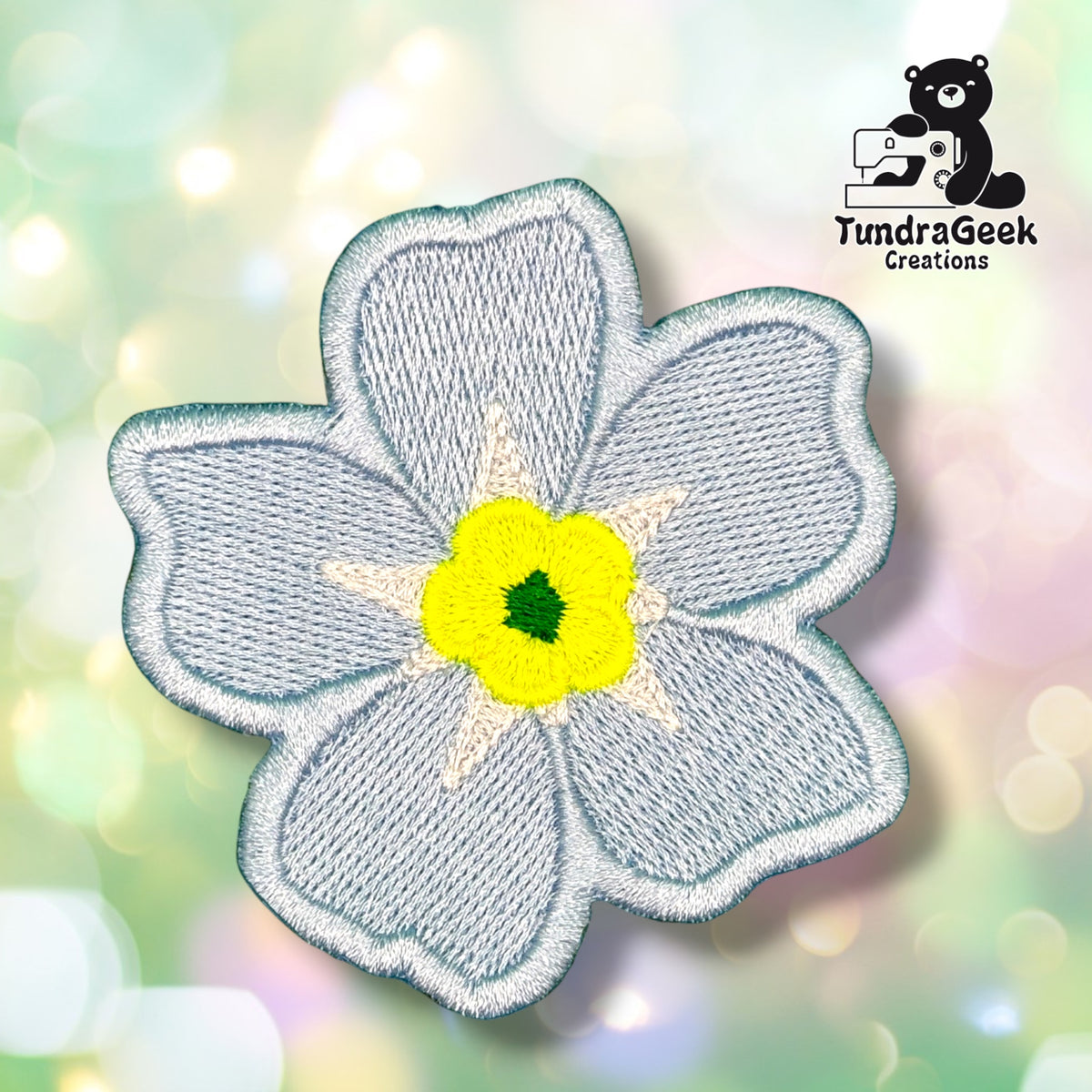 Front view of a five-petaled flower-shaped patch embroidered in soft grey-blue with a yellow center and small green accent, with a white star-shaped ring around the center.