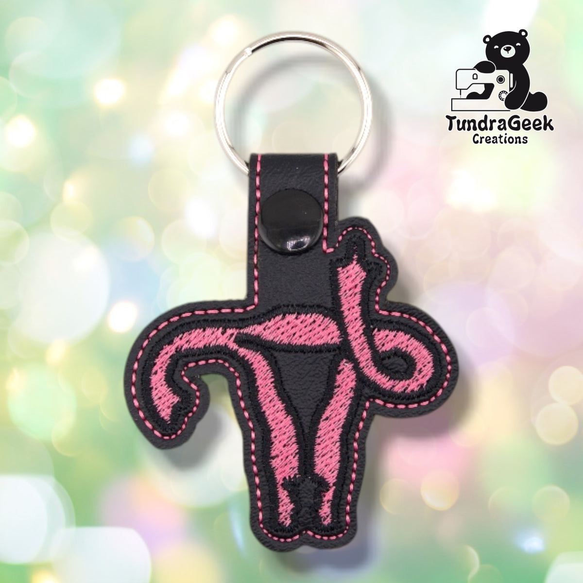 Black vinyl keychain shaped around a uterus with one fallopian tube curling upward into a raised middle finger. The design is filled in pink satin stitch with a black outline, with pink topstitching around the edge and up the snap tab. A black plastic snap closes the tab around a silver split ring.