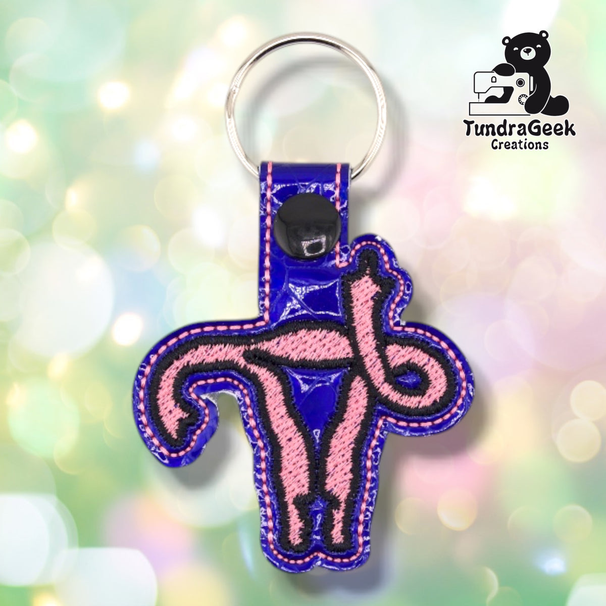 Metallic Deep Blue colorway in textured patent vinyl with a crackled sheen, carrying the same uterus design in pink satin stitch with a black outline. Pink topstitching runs around the edge, and a black plastic snap closes the tab.
