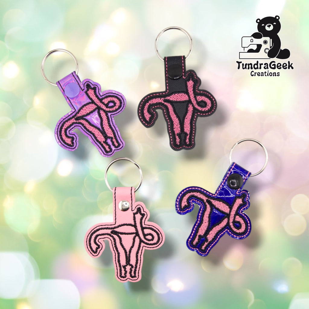 Four F Uterus keychains arranged together, showing all colorways: iridescent lavender with a lavender snap, smooth black with a black snap, smooth pink with a silver rivet, and textured deep blue patent with a black snap. Three carry the uterus design in pink satin stitch outlined in black; the pink one is stitched in black outline only.