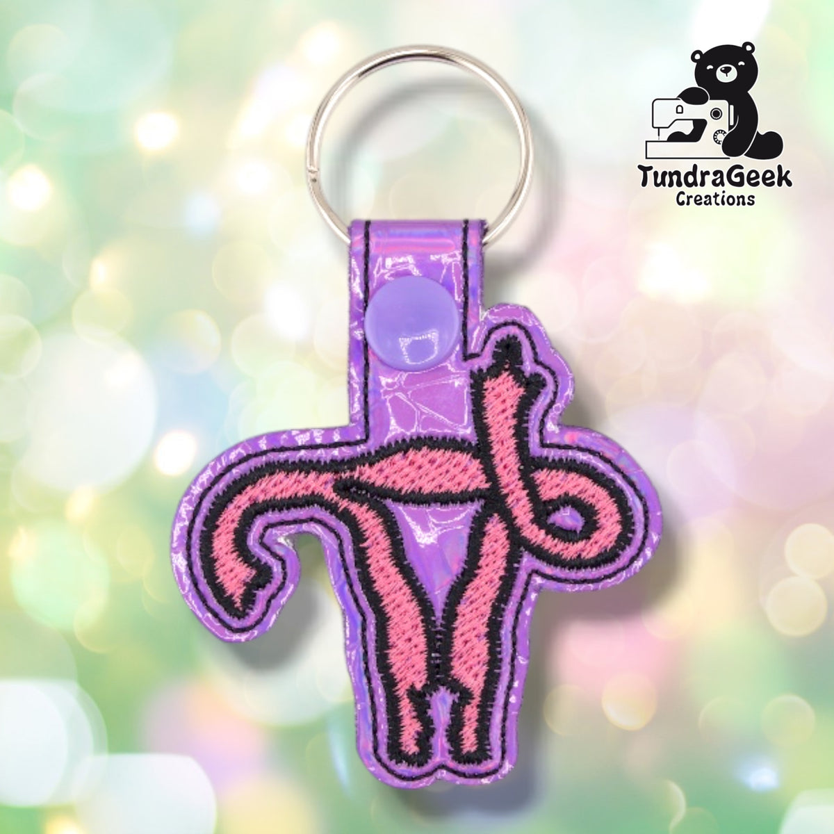 Metallic Lavender colorway in iridescent vinyl that shifts between lavender and pink, with the uterus design in pink satin stitch outlined in black. A lavender plastic snap closes the tab around a silver split ring.