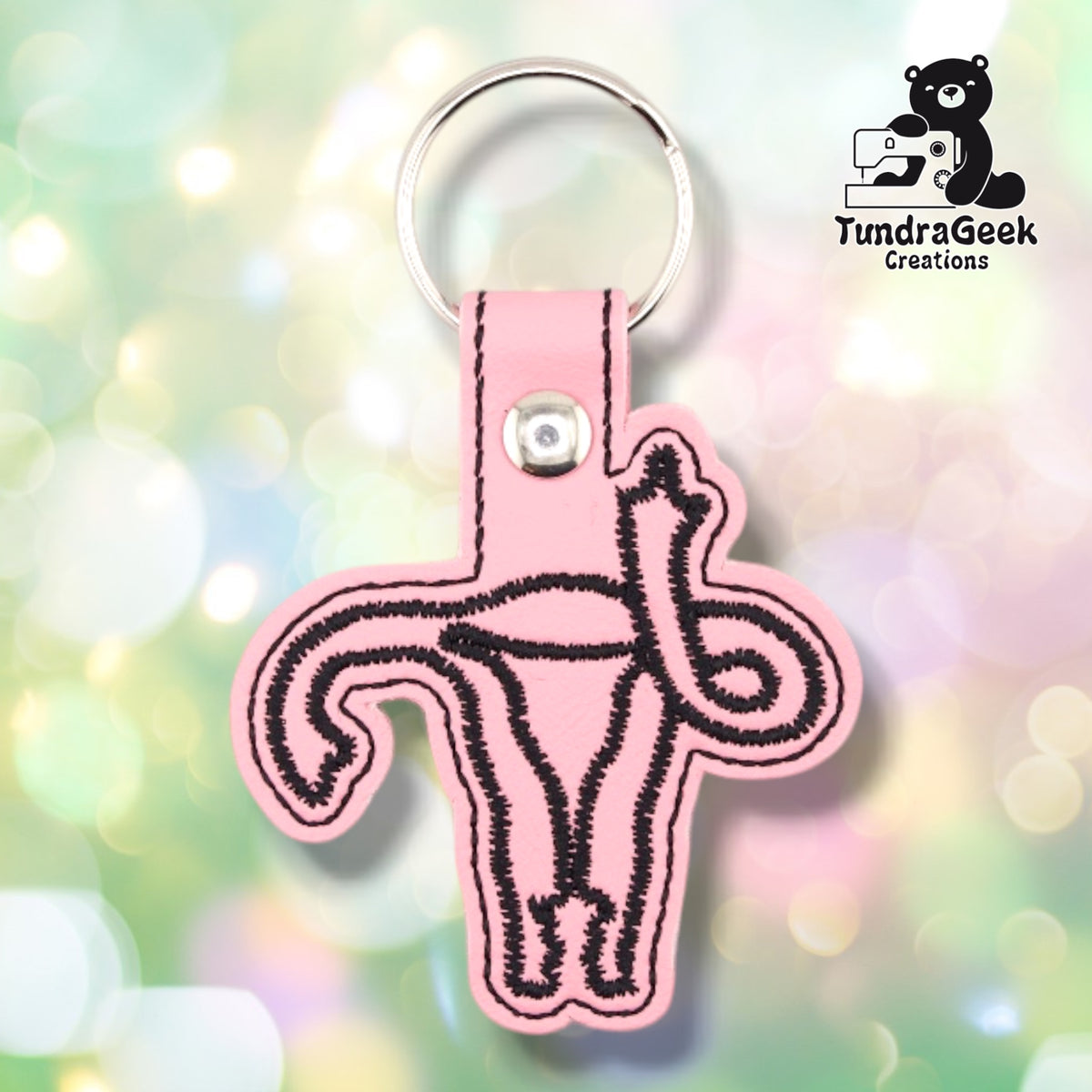 Pink colorway in smooth pink vinyl. The uterus design is stitched in black outline only, with no fill, so the pink vinyl shows through the shape. A silver metal rivet closes the tab around a silver split ring.