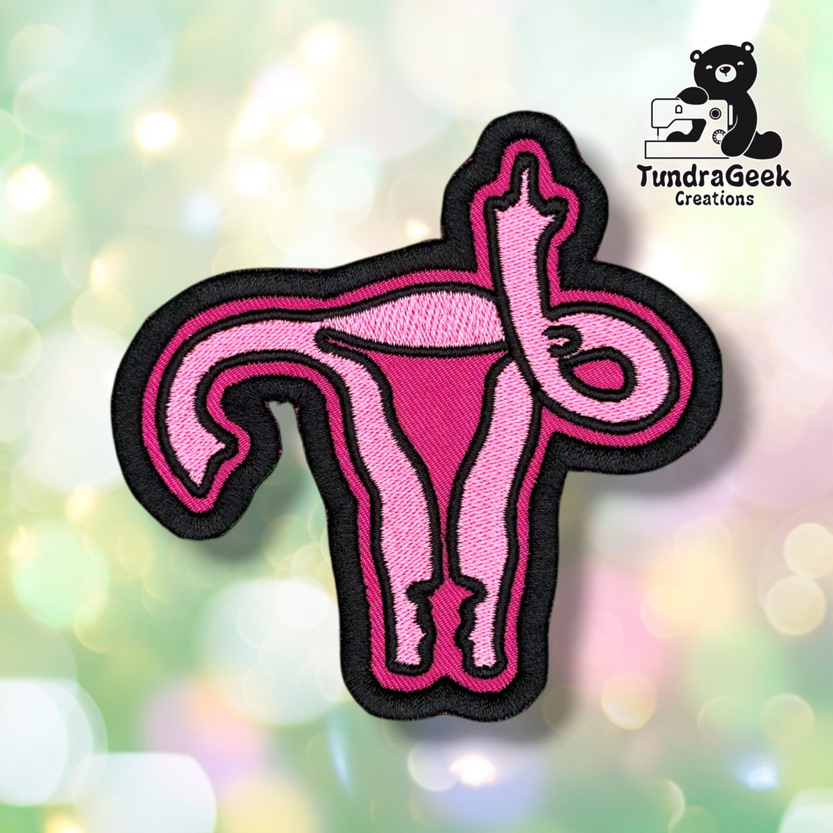 Front view of a patch shaped like a uterus and fallopian tubes outlined in black, embroidered in shades of pink, with the left fallopian tube and ovary stylized into a raised middle finger.
