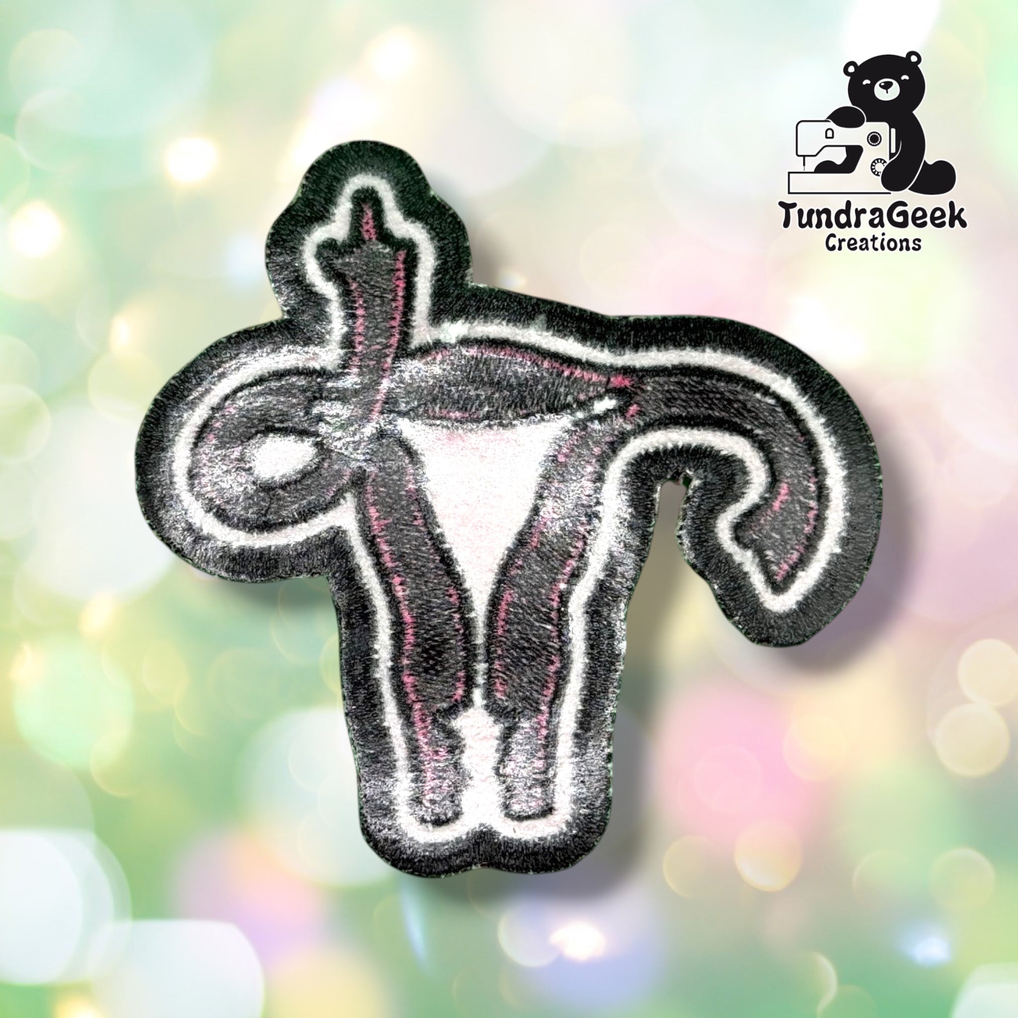 Back of the patch showing the reverse side of the embroidery in muted grey, white, and pink thread, with the uterus and raised-finger shape faintly visible through the backing.