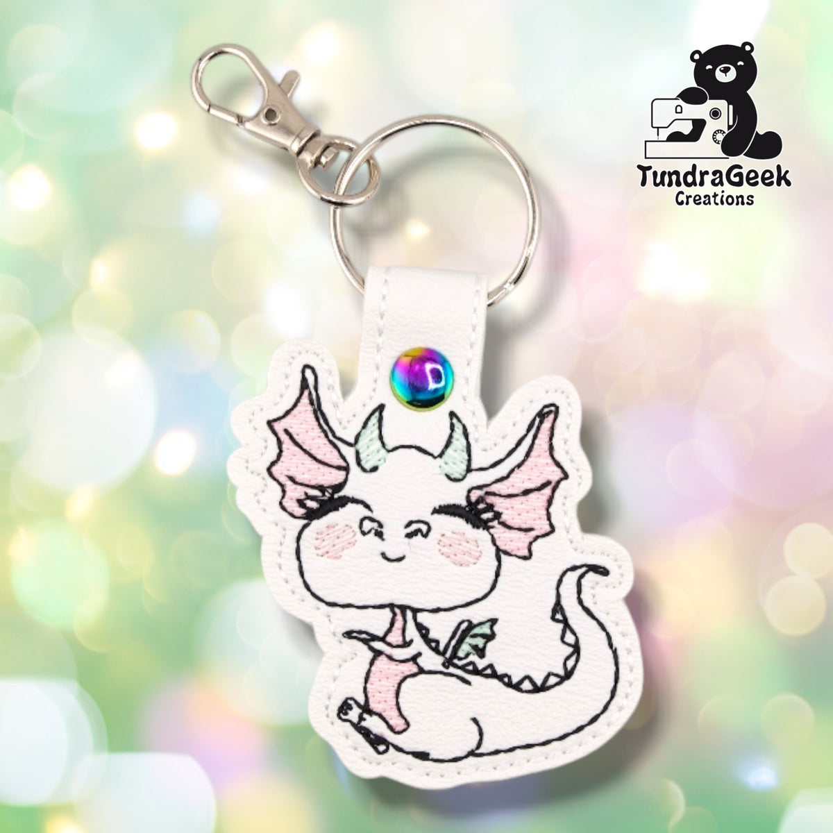 White faux leather keychain shaped around a curled-up cartoon dragon, embroidered in fine black outline with soft pink wings and blushed cheeks and pale mint horns and back spines. The dragon's eyes are closed in a contented smile and her tail curls around the base. The snap tab at the top is closed with an iridescent rainbow snap and carries a split ring and a silver swivel lobster clasp.