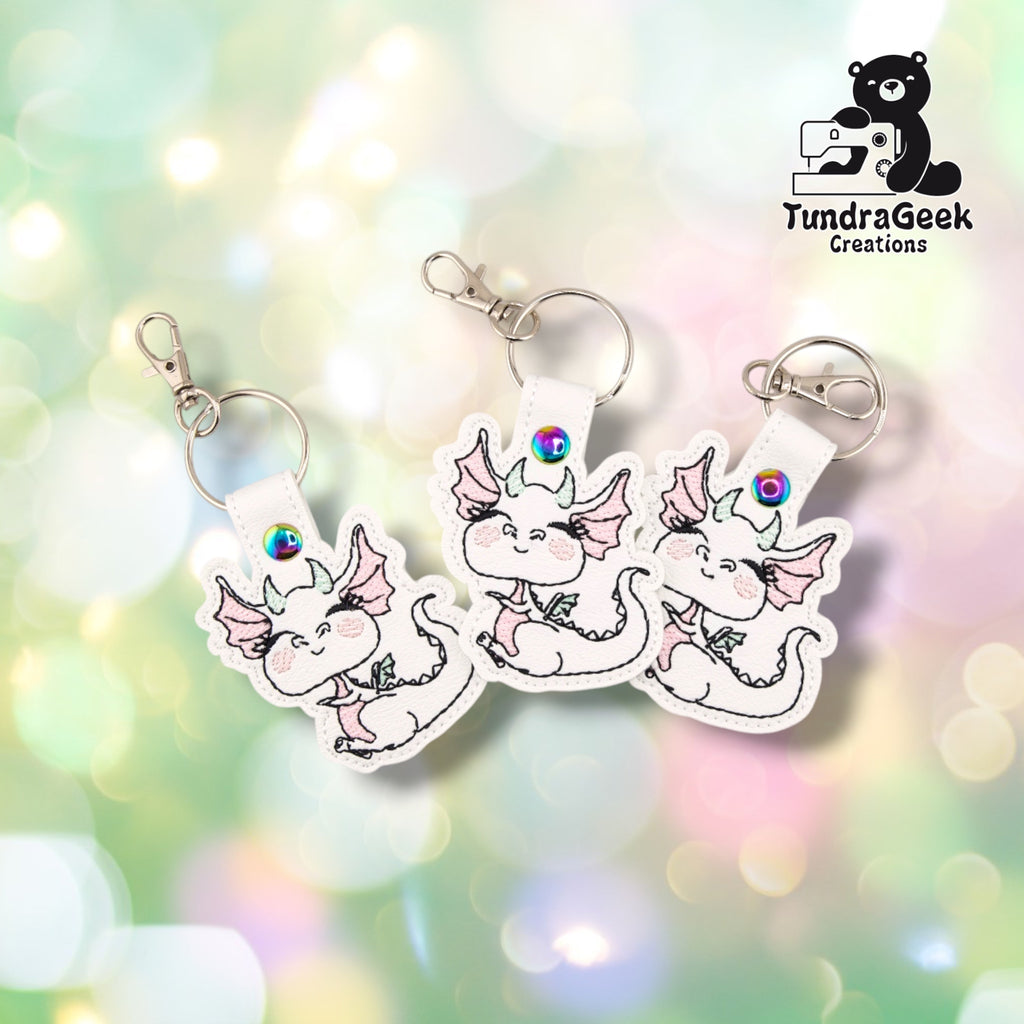 Three identical Femme Dragon keychains arranged at slightly different angles, each on white faux leather with the same pink and mint line-art dragon, iridescent snap, split ring and silver swivel lobster clasp.
