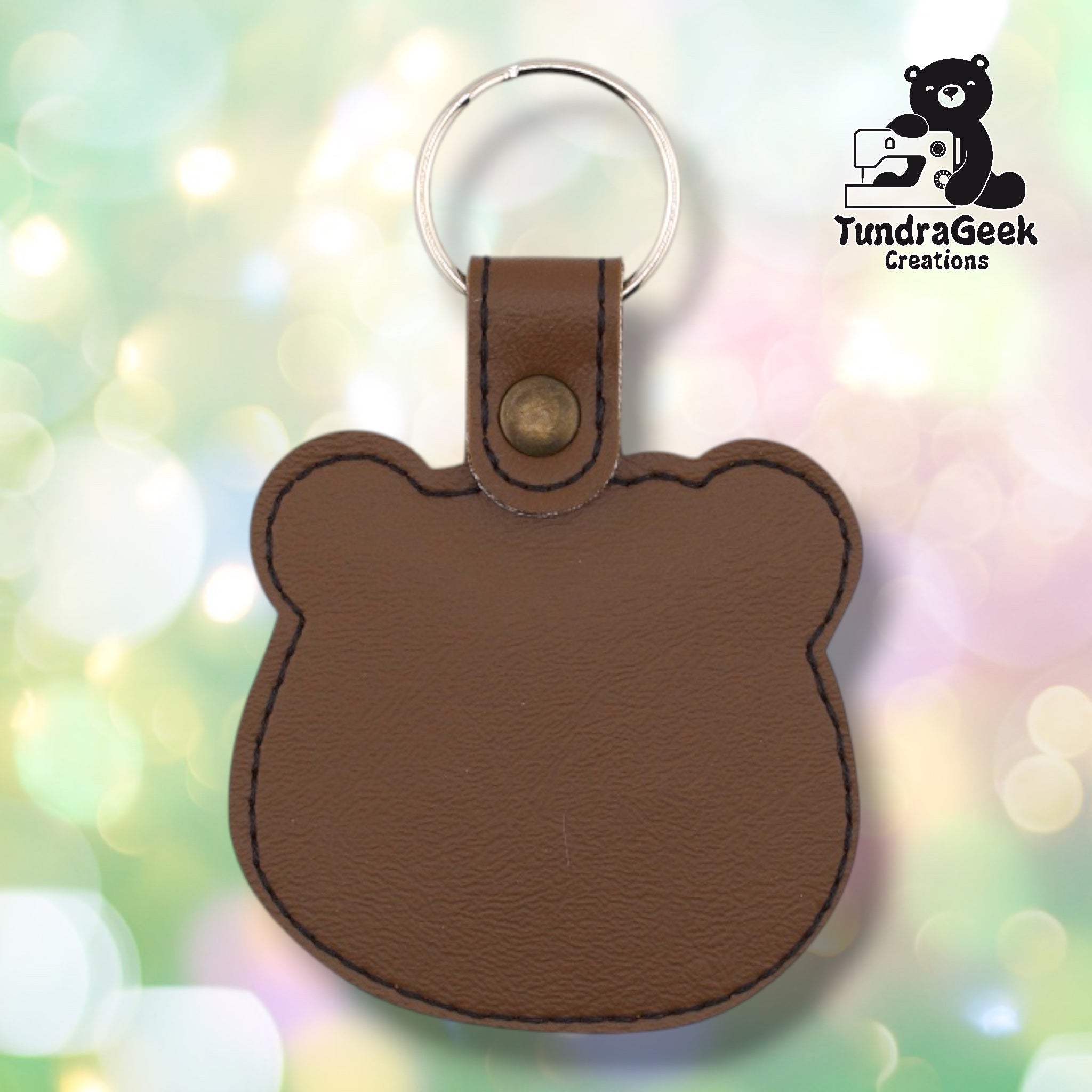 Back of a brown faux leather floral bear keychain. The plain brown vinyl back panel shows dark outline stitching following the rounded bear head silhouette shape. A bronze 9mm metal rivet closure secures the tab, with a silver split ring attached above.