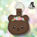 Front of a brown faux leather floral bear keychain. An embroidered bear face features large dark eyes with white highlights, a small black nose, and a subtle smile on a brown vinyl base. A flower crown of pink, white, and lavender blooms with yellow centers and green leaves sits across the top of the bear's head, with brown ear accents on either side. A bronze 9mm metal rivet closure is visible on the tab, with a silver split ring above.