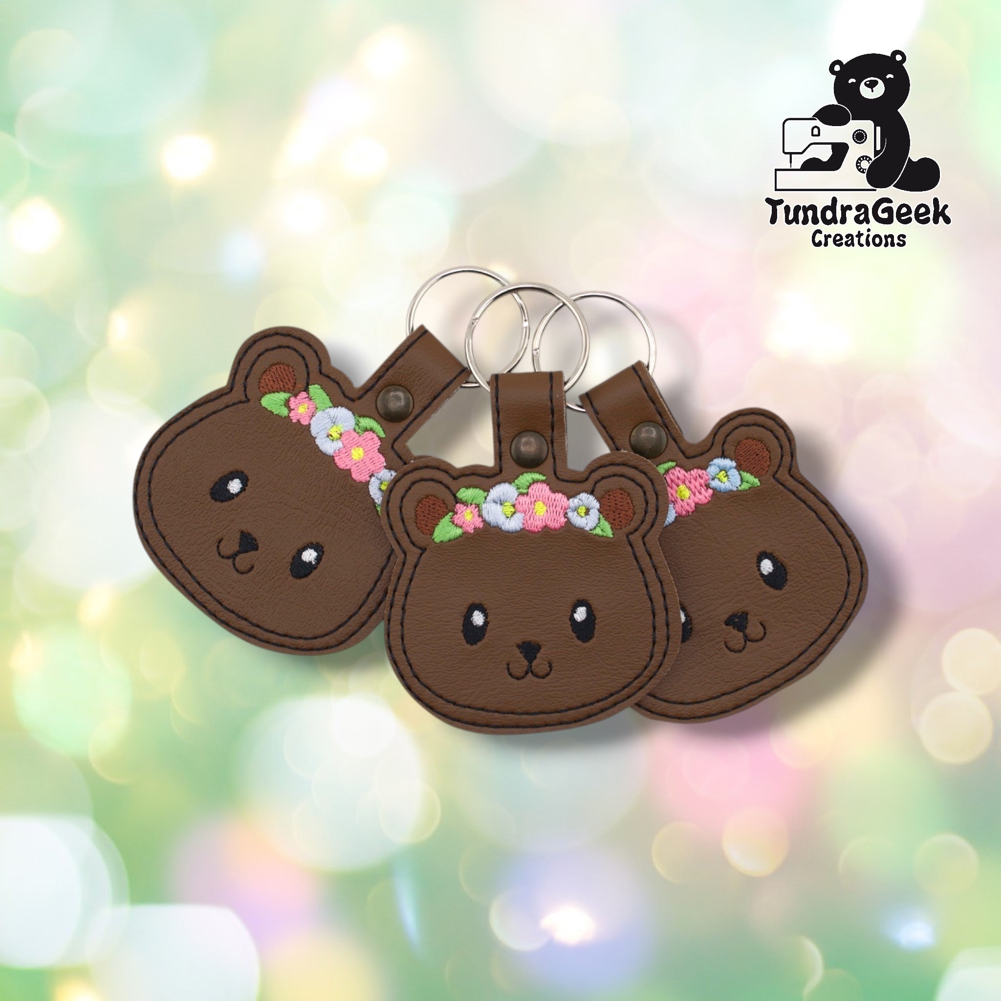Three floral bear keychains arranged together against a soft bokeh background, all on brown vinyl. Slight variation in flower crown thread colors is visible across the three, illustrating natural small-batch variation in the embroidery fill.