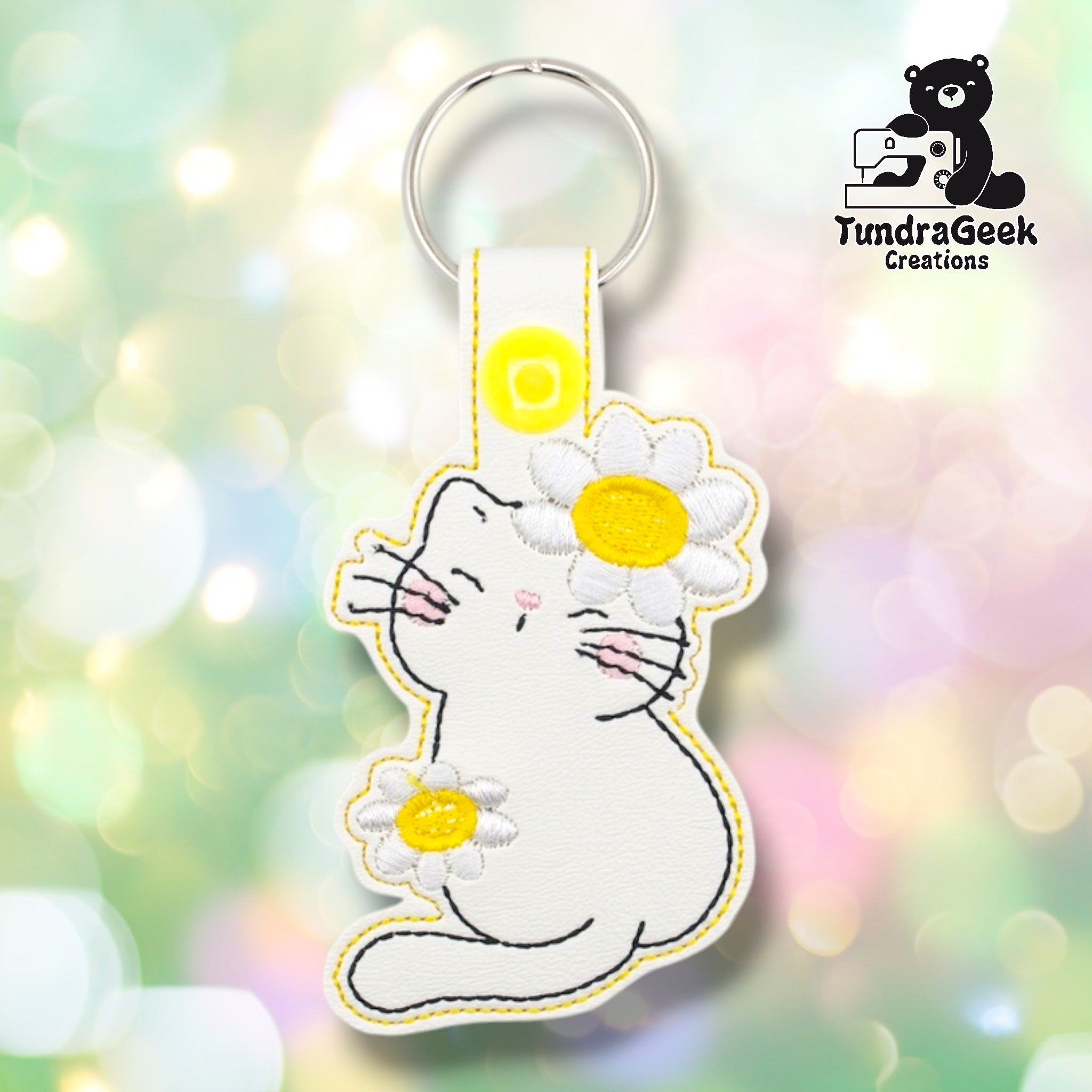 White faux leather keychain shaped like a chubby sitting cat holding daisies. A large daisy with white petals and yellow center rests against the cat's face; a smaller daisy is held in one paw. Black embroidery details the cat's features; pink blush marks the cheeks and inner ears. Yellow plastic snap closure and silver key ring at top. TundraGeek Creations logo in upper right corner.