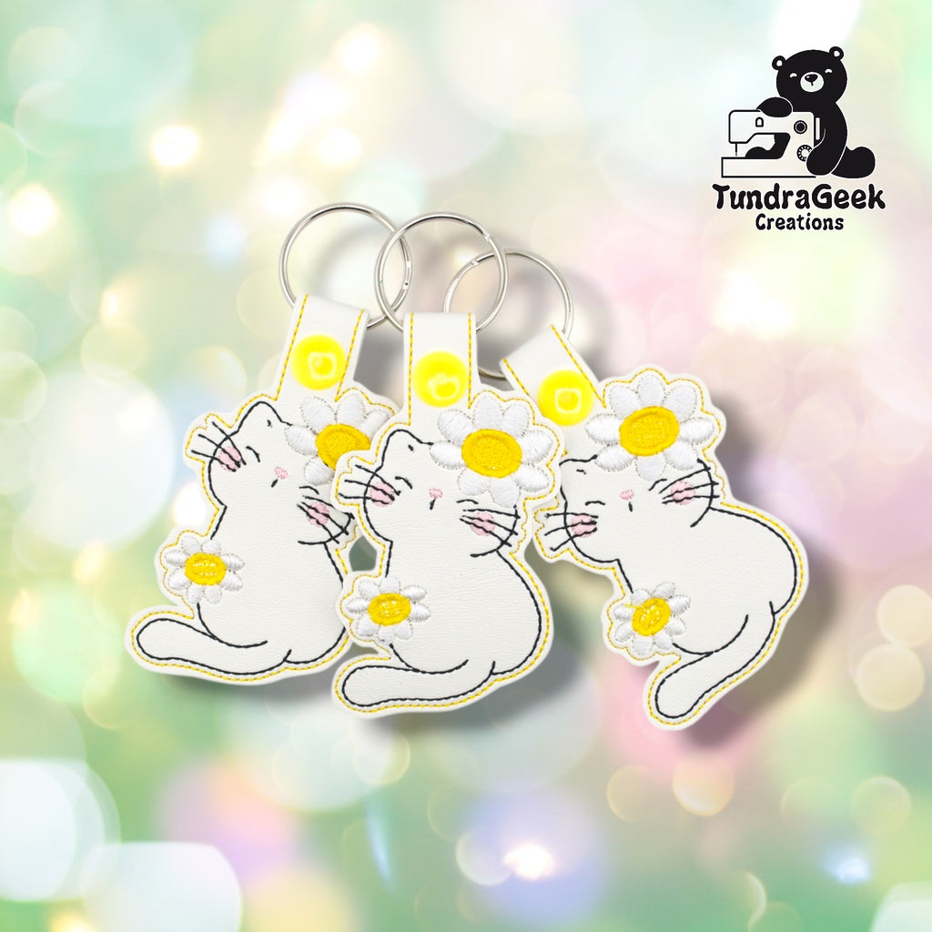 Three flower kitty keychains arranged together on a pastel bokeh background, showing the design from slightly varied angles. All three feature matching white faux leather, daisy embroidery, and yellow snap closures.