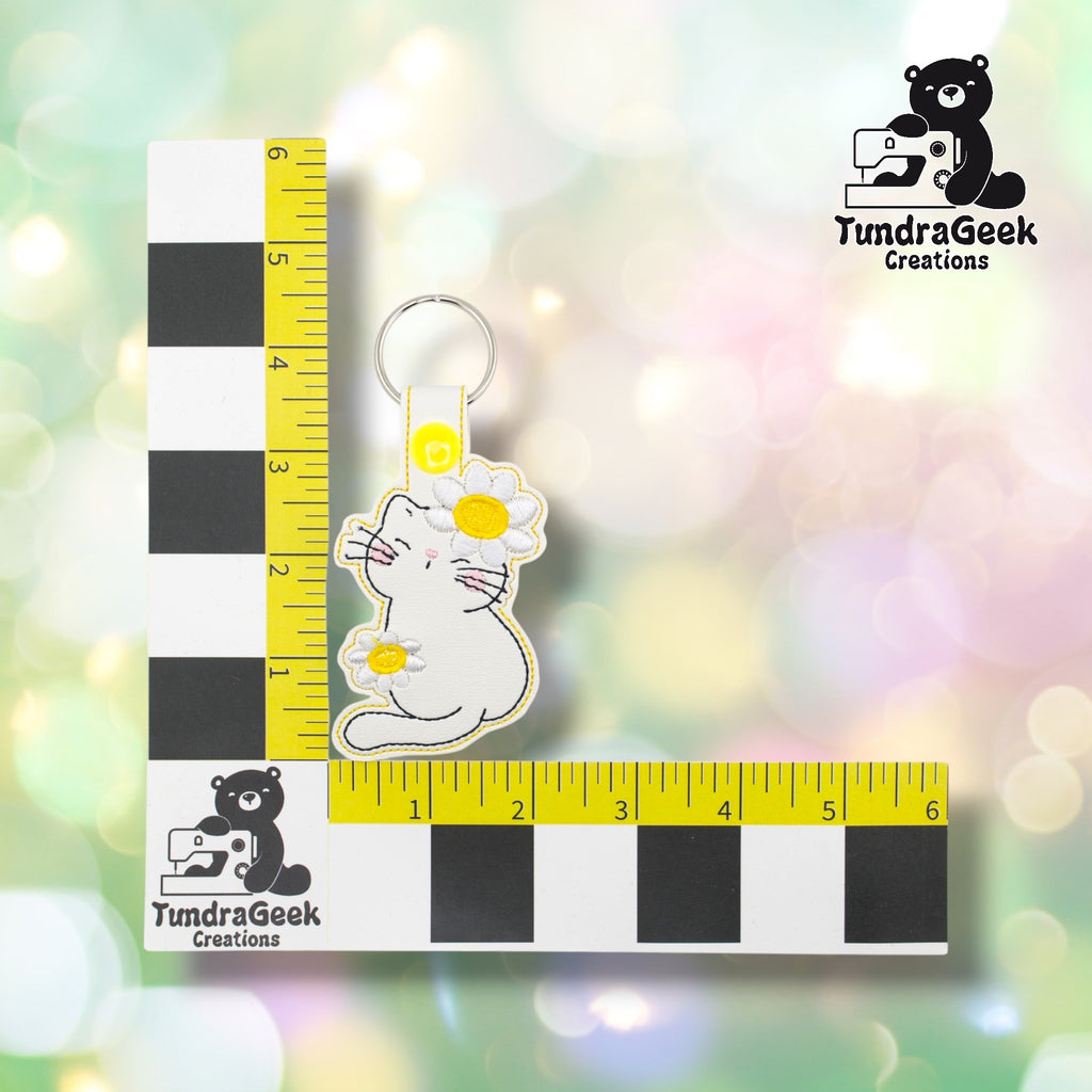 Flower kitty keychain positioned against a TundraGeek Creations branded ruler card showing yellow tape measure. Keychain body measures approximately 2.5 inches wide by 3 inches tall, not including hardware.