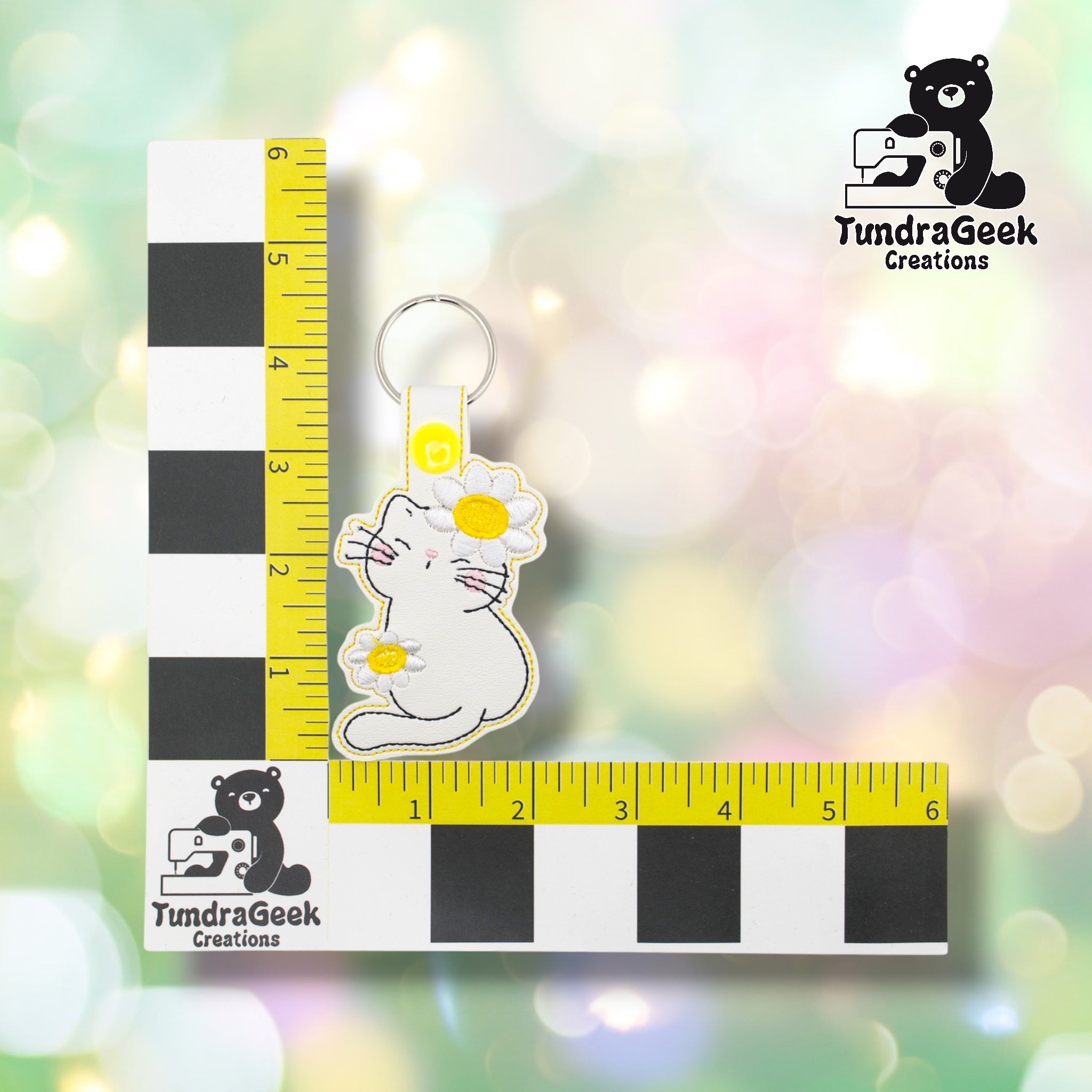 Flower kitty keychain positioned against a TundraGeek Creations branded ruler card showing yellow tape measure. Keychain body measures approximately 2.5 inches wide by 3 inches tall, not including hardware.
