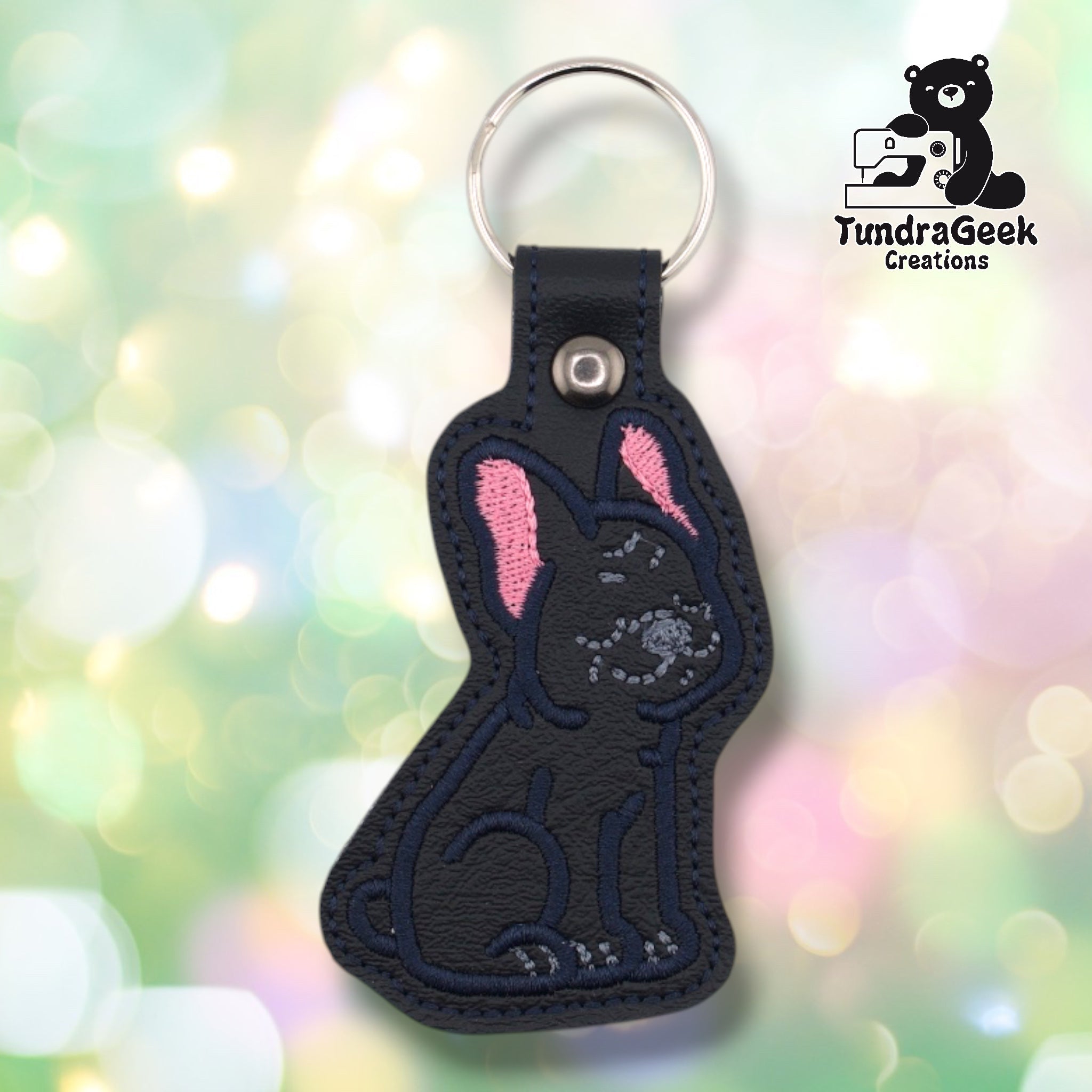 Front of a black faux leather French Bulldog keychain. An embroidered French Bulldog sits upright on a black vinyl base with pink inner ear accents, navy outline stitching, and gray facial detail stitching capturing the breed's characteristic wrinkles and expression. A gunmetal 9mm metal rivet closure is visible on the tab, with a silver split ring above.