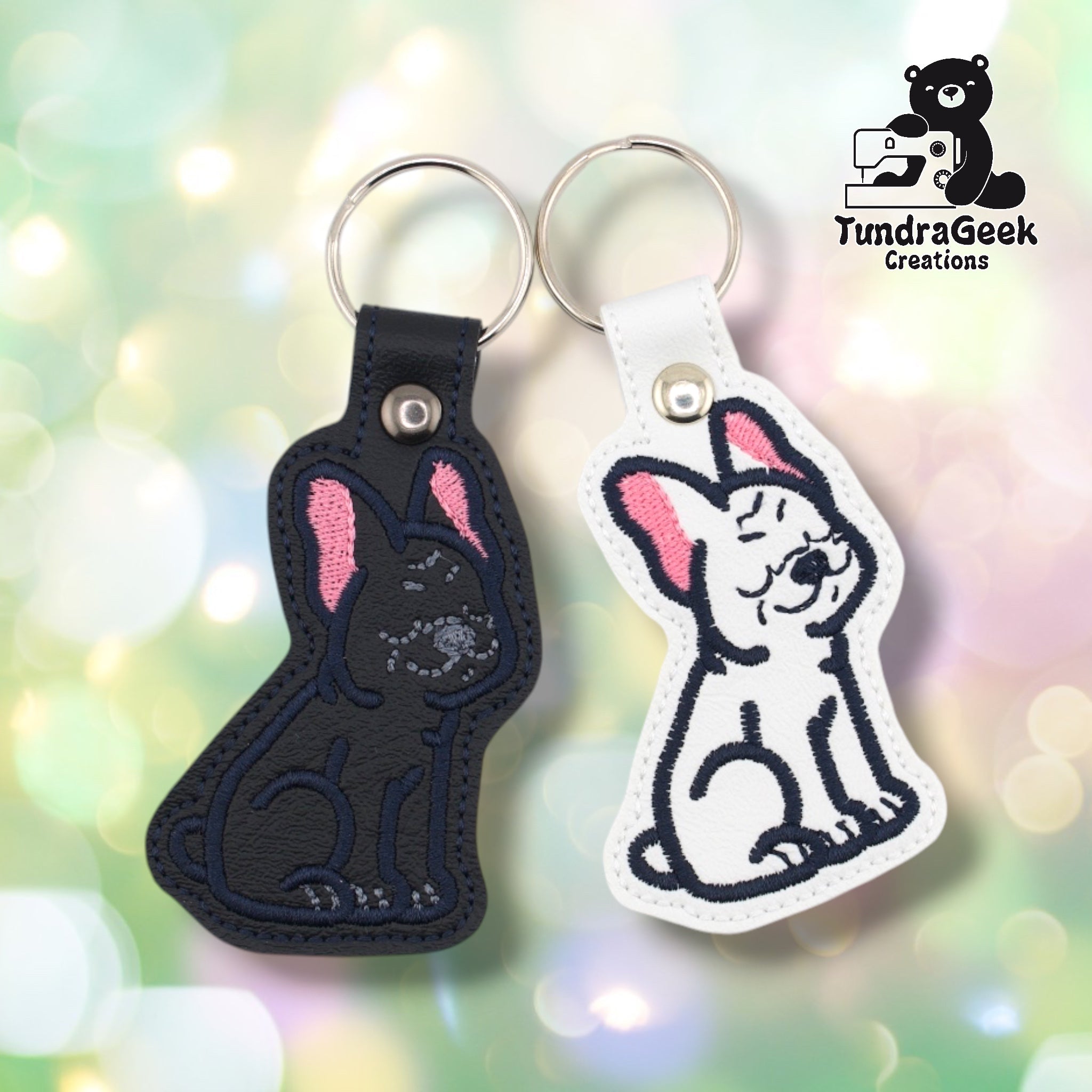 Both French Bulldog keychain variants shown side by side against a soft bokeh background. Black variant on the left, white variant on the right. Both feature the same embroidered Frenchie design with pink inner ear accents.