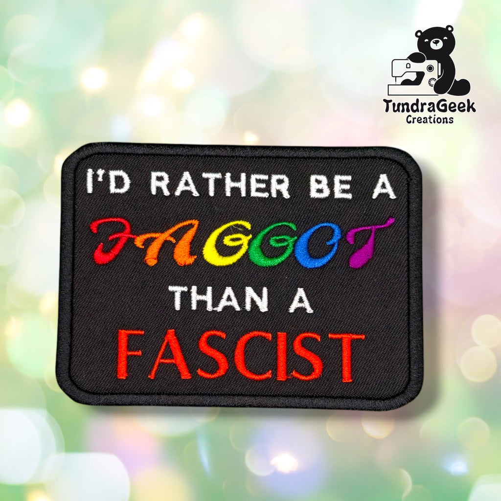 Front view of a black rectangular patch with a black merrowed edge, reading "I'D RATHER BE A" in white, a reclaimed anti-gay slur in rainbow lettering, "THAN A" in white, and "FASCIST" in bold red block letters.