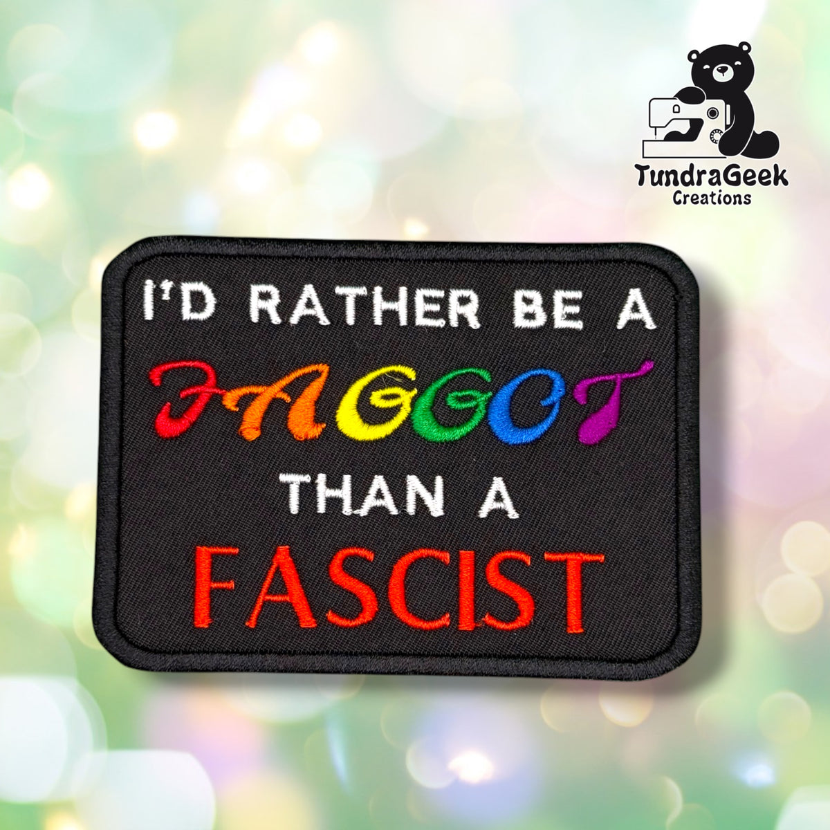 Front view of a black rectangular patch with a black merrowed edge, reading "I'D RATHER BE A" in white, a reclaimed anti-gay slur in rainbow lettering, "THAN A" in white, and "FASCIST" in bold red block letters.
