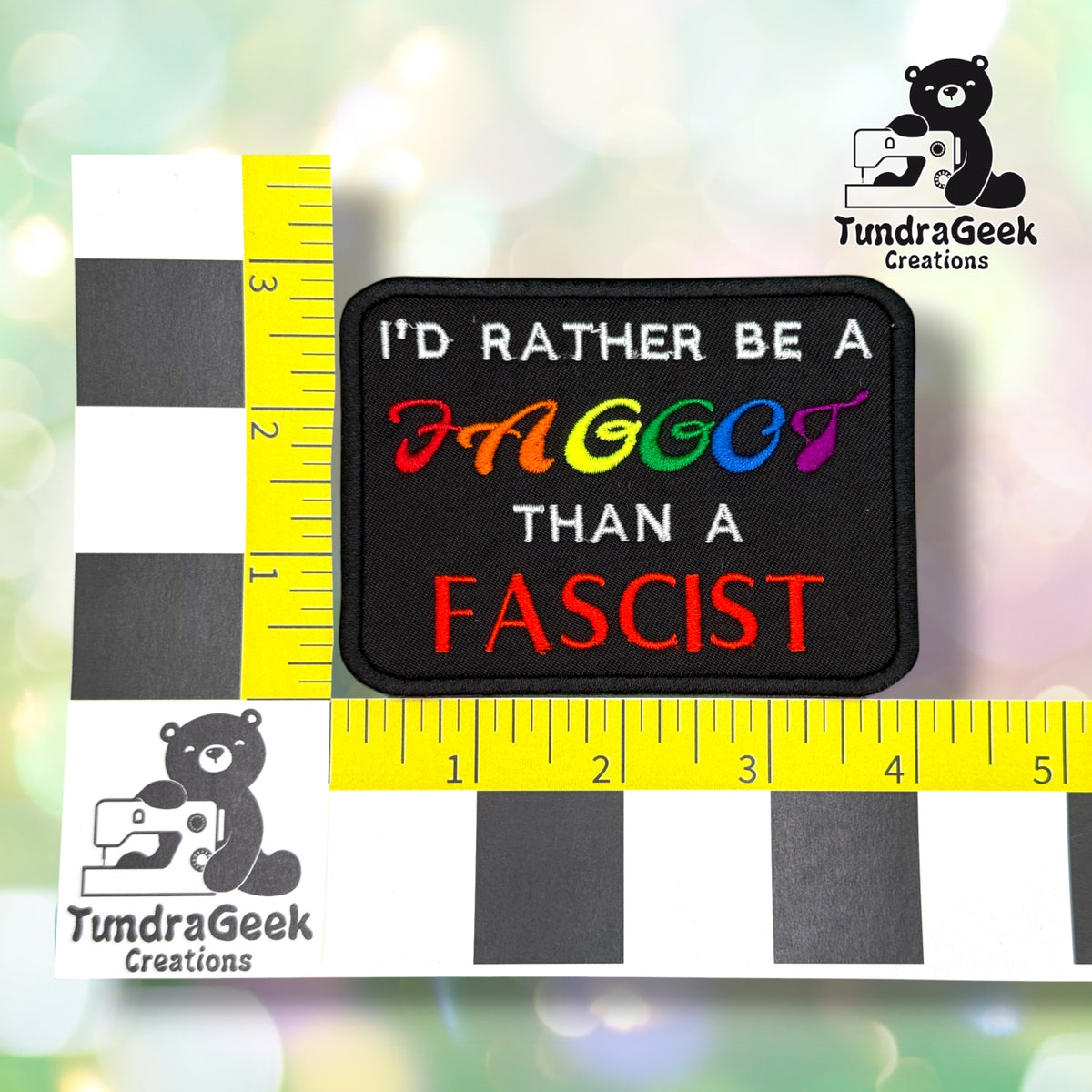 Patch shown against a black-and-white checkered background with yellow rulers for scale, measuring approximately 4 inches wide by 2.75 inches tall.