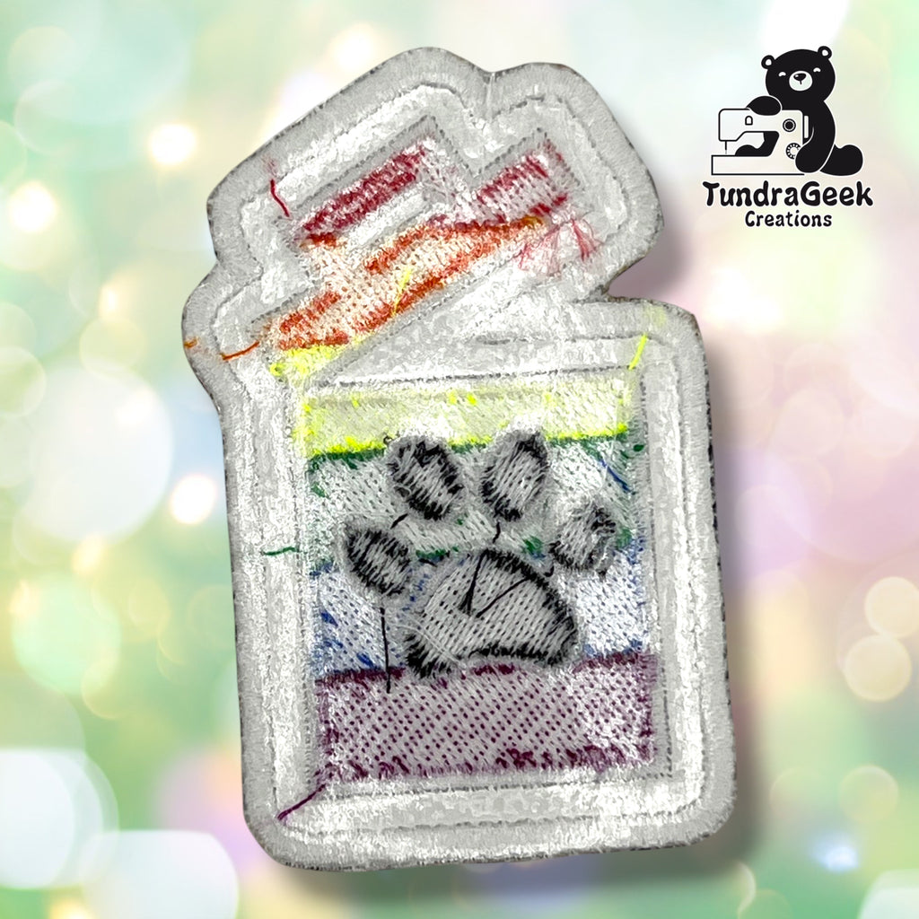 The backside of a small embroidered patch shaped like a trash can with a rainbow stripe pattern and a black paw print on the front. 