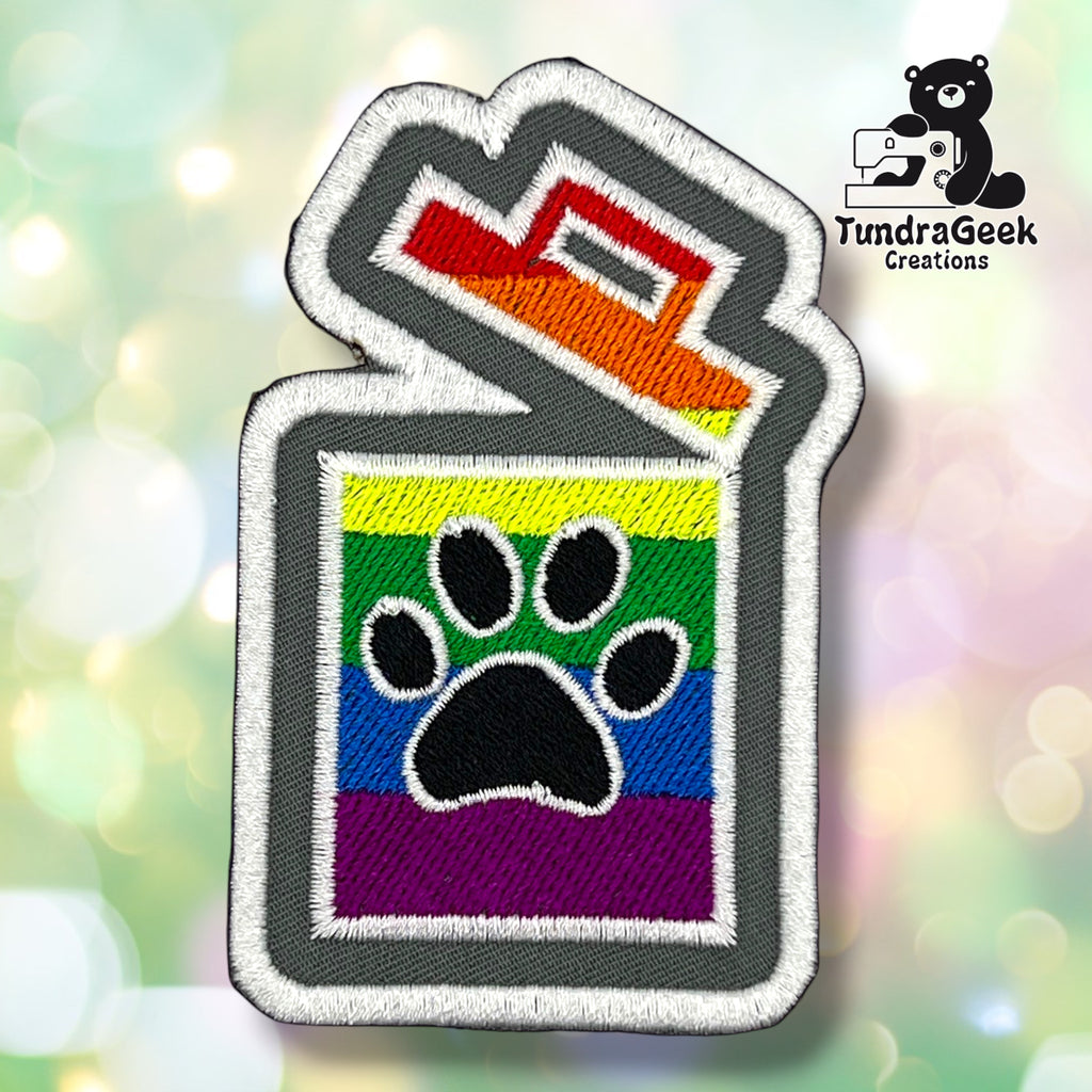 An embroidered patch shaped like a trash can with a paw print, featuring a rainbow stripe pattern in yellow, green, blue, purple, and red, with a gray border and white stitching. The patch is shown against a soft, blurred multicolor bokeh background.