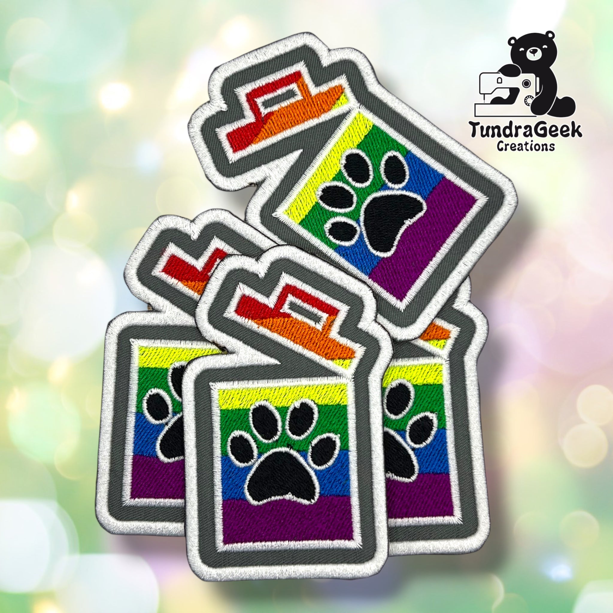Four embroidered patches with a rainbow stripe background and a black paw print in the center, arranged on a soft pastel bokeh background. A TundraGeek Creations logo is visible in the top right.