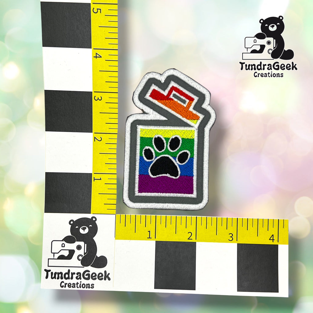 A small embroidered patch shaped like a trash can with a rainbow stripe pattern and a black paw print on the front, shown with measuring rulers and TundraGeek Creations logos on a soft blurred pastel background.
