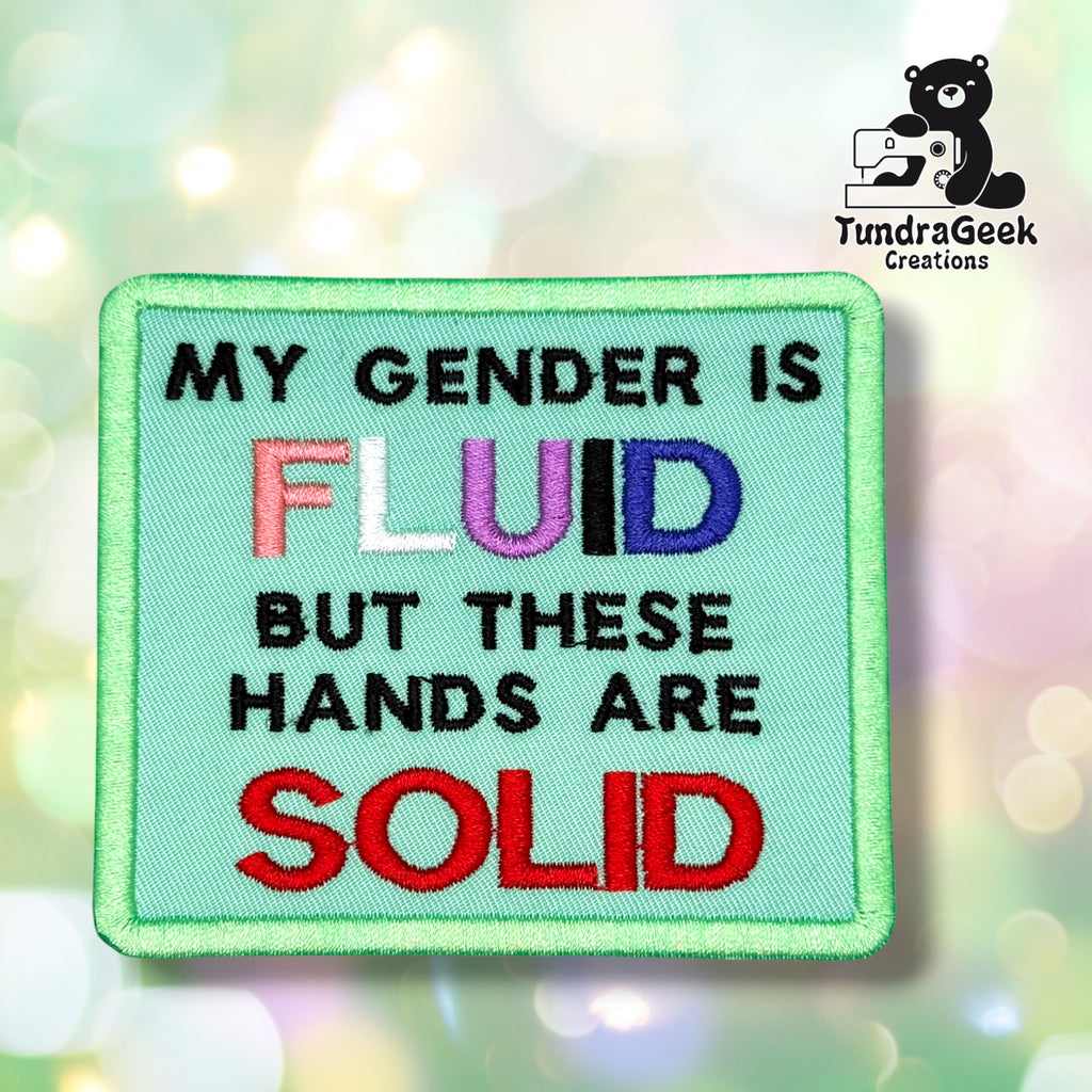 Front view of a mint green rectangular patch with a green merrowed edge, reading "MY GENDER IS" in black, "FLUID" in genderfluid pride flag colors (pink, white, purple, black, blue), "BUT THESE HANDS ARE" in black, and "SOLID" in red.