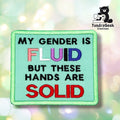 Front view of a mint green rectangular patch with a green merrowed edge, reading "MY GENDER IS" in black, "FLUID" in genderfluid pride flag colors (pink, white, purple, black, blue), "BUT THESE HANDS ARE" in black, and "SOLID" in red.