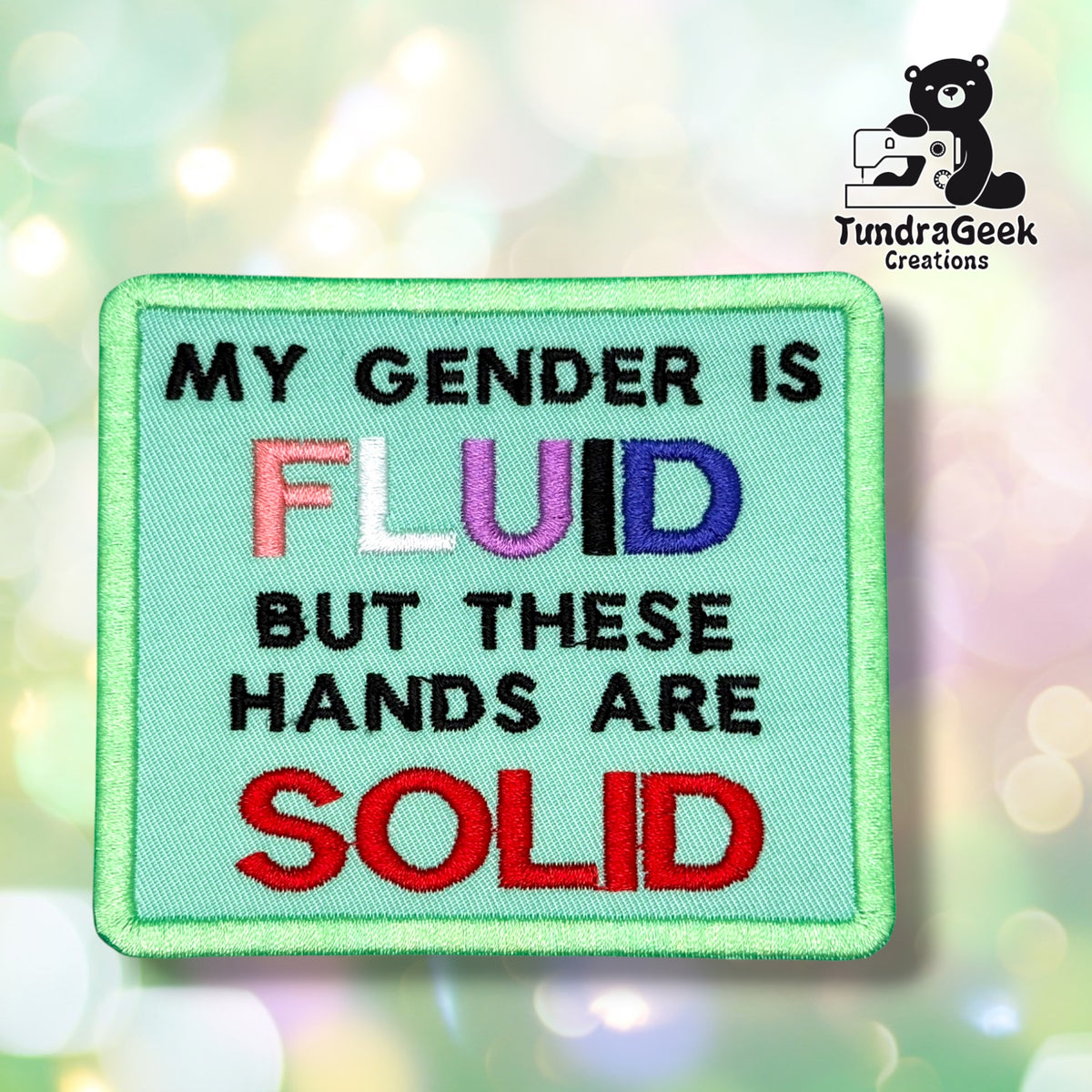 Front view of a mint green rectangular patch with a green merrowed edge, reading "MY GENDER IS" in black, "FLUID" in genderfluid pride flag colors (pink, white, purple, black, blue), "BUT THESE HANDS ARE" in black, and "SOLID" in red.