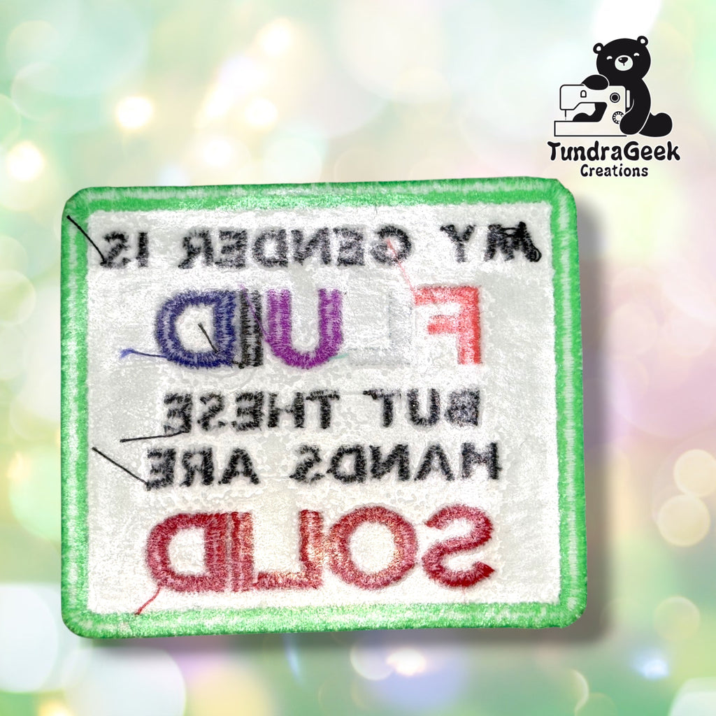 Back of the patch showing the reverse side of the embroidery in white and pastel thread, with the mirrored lettering faintly visible through the backing.