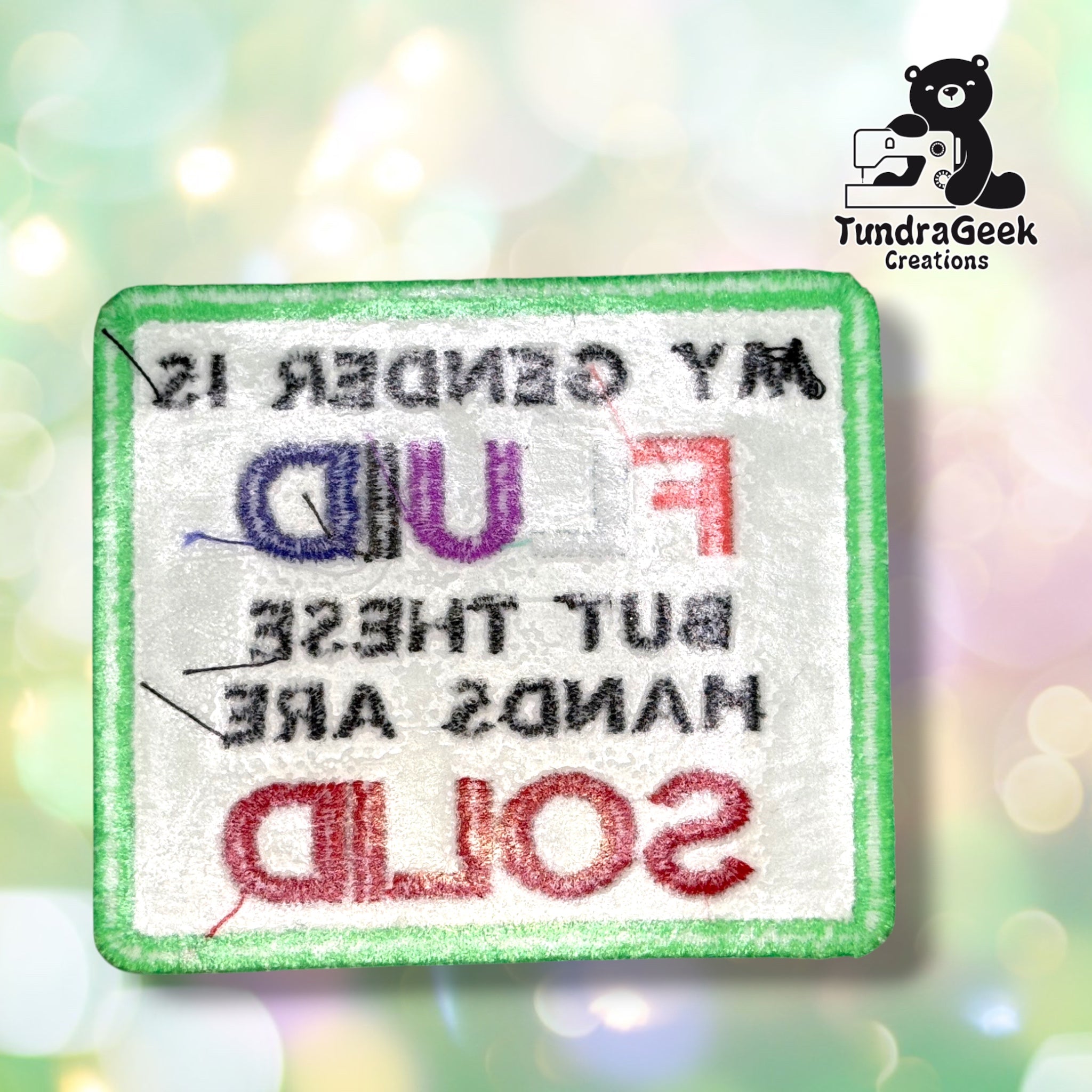 Back of the patch showing the reverse side of the embroidery in white and pastel thread, with the mirrored lettering faintly visible through the backing.