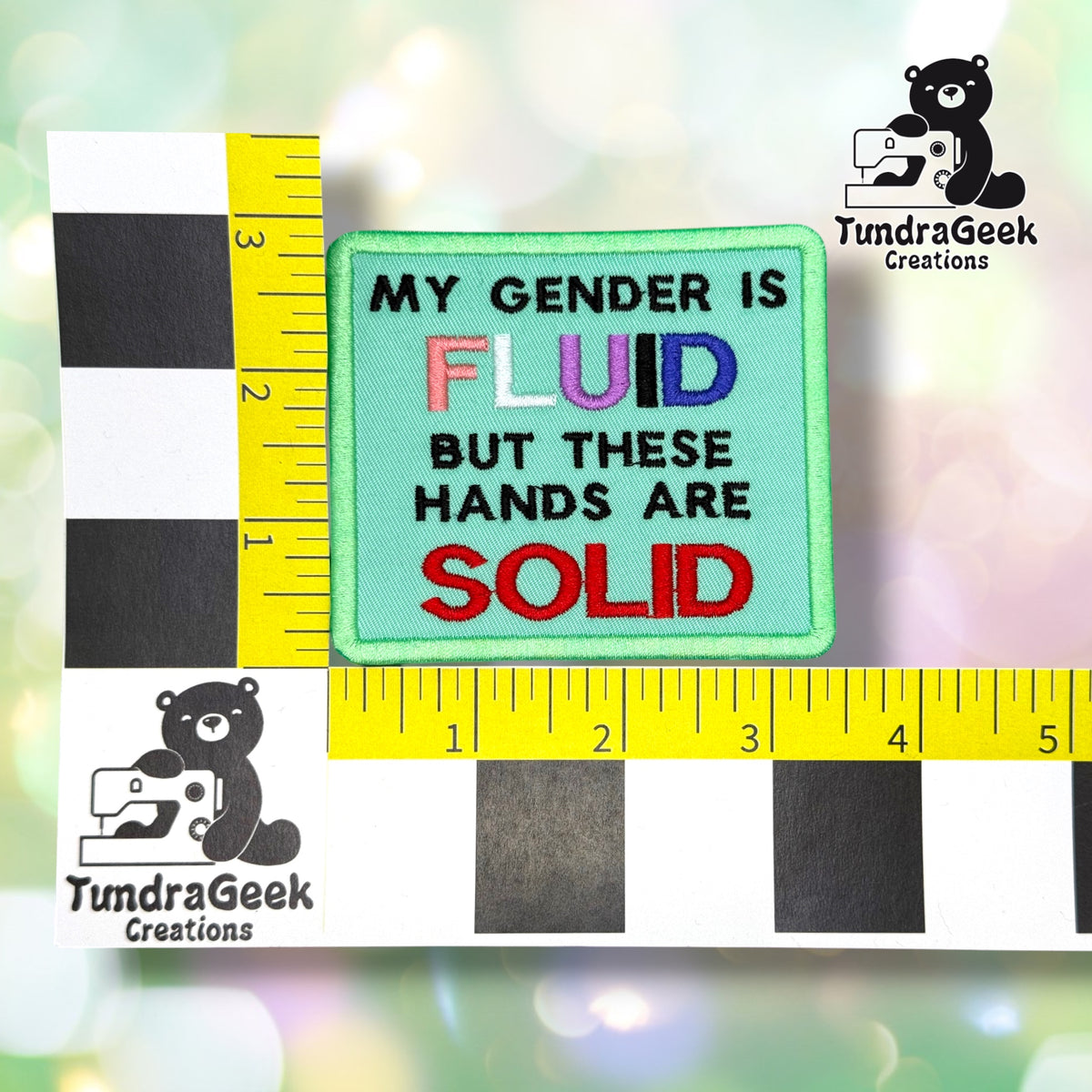 Patch shown against a black-and-white checkered background with yellow rulers for scale, measuring approximately 3.5 inches wide by 4 inches tall.  ---