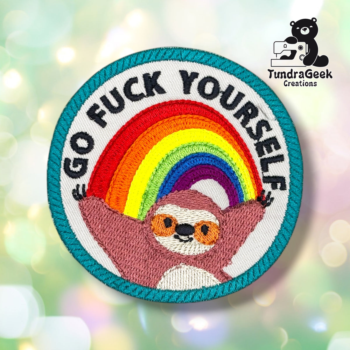 Front view of a circular patch with a teal merrowed edge, a rude four-word phrase embroidered in black block letters arcing around a rainbow (red, orange, yellow, green, blue, purple), with a smiling rose-pink sloth hanging from the rainbow's ends, cream face and belly, and orange-ringed eyes.