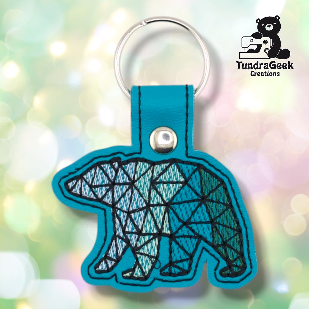 Front of a teal faux leather geometric polar bear keychain. An embroidered low-poly polar bear design fills the bear-shaped vinyl base, with thread color transitioning from light blue and white at the head and shoulders to teal and green toward the hindquarters. Black geometric grid lines define the faceted shapes throughout. A silver 9mm metal rivet closure is visible on the tab, with a silver split ring above.