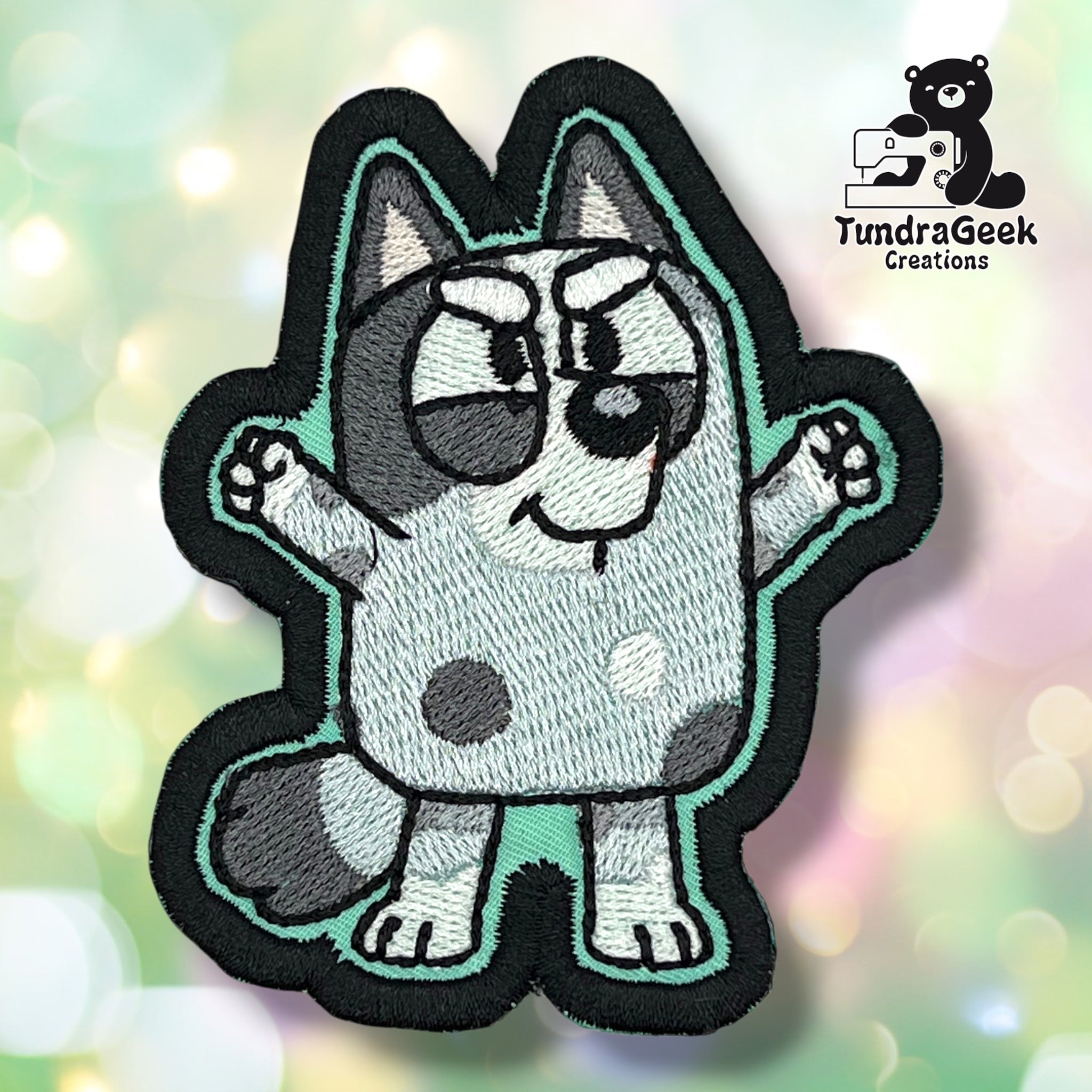 An embroidered patch of a cartoon gray and white dog with raised arms, black outlines, and a confident expression. TundraGeek Creations logo is in the top left corner.