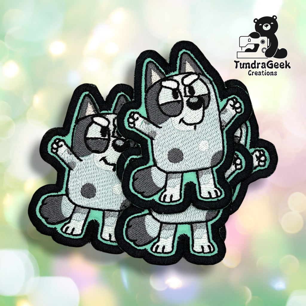 Three embroidered patches featuring a cartoon blue dog with spots and raised arms, displayed on a geometric light background. TundraGeek Creations logo in the top left corner.