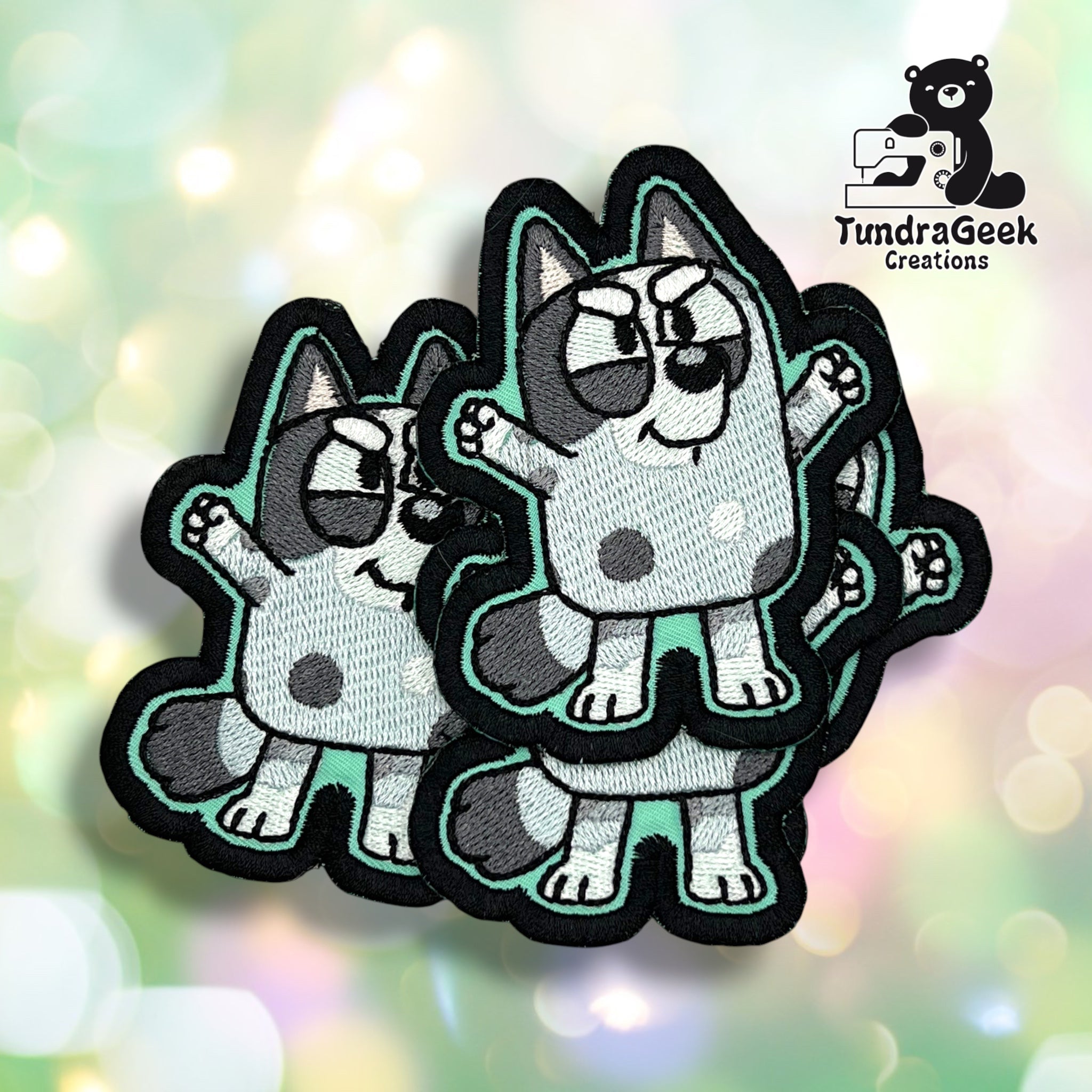 Three embroidered patches featuring a cartoon blue dog with spots and raised arms, displayed on a geometric light background. TundraGeek Creations logo in the top left corner.