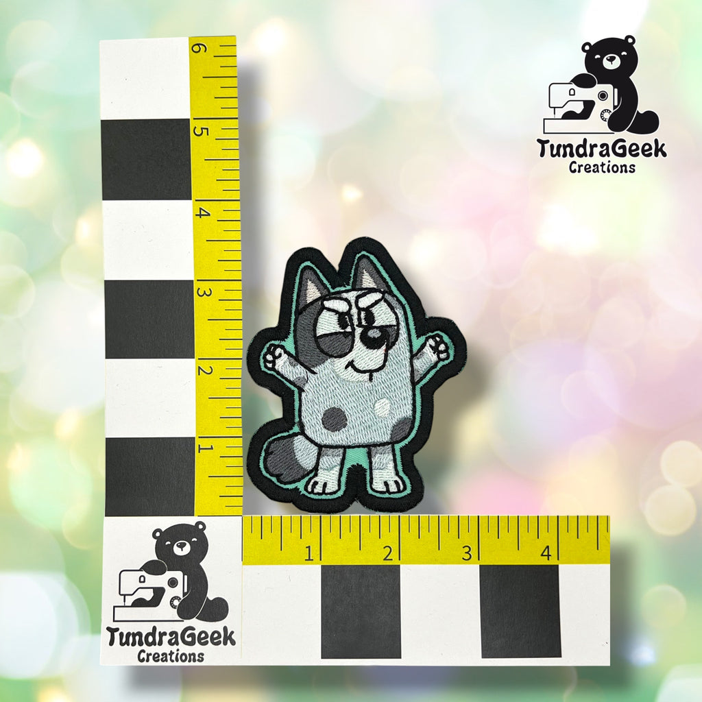 A small embroidered patch of a cartoon dog is placed on a checkered measuring grid with TundraGeek Creations logos on a pastel bokeh-style background.