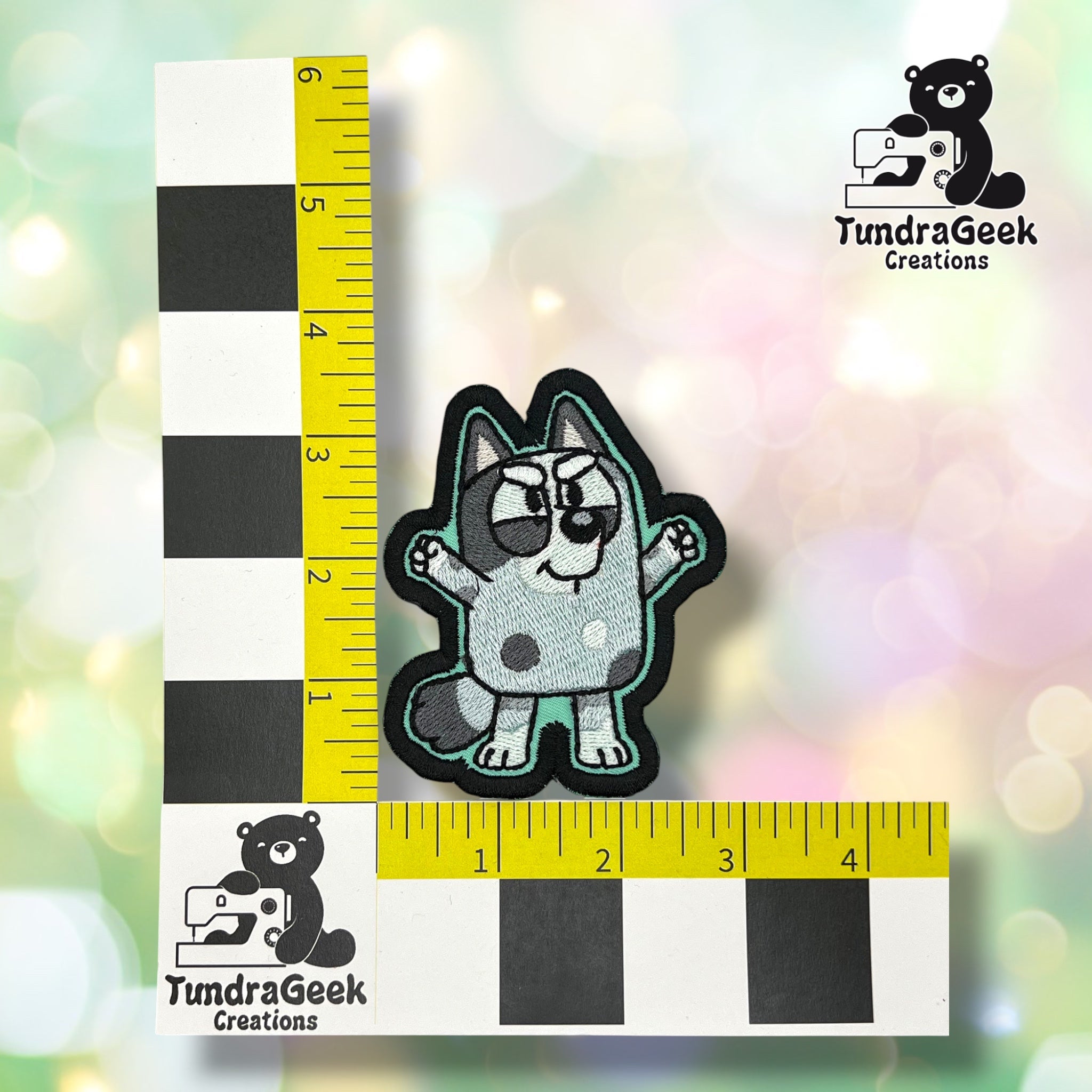 A small embroidered patch of a cartoon dog is placed on a checkered measuring grid with TundraGeek Creations logos on a pastel bokeh-style background.