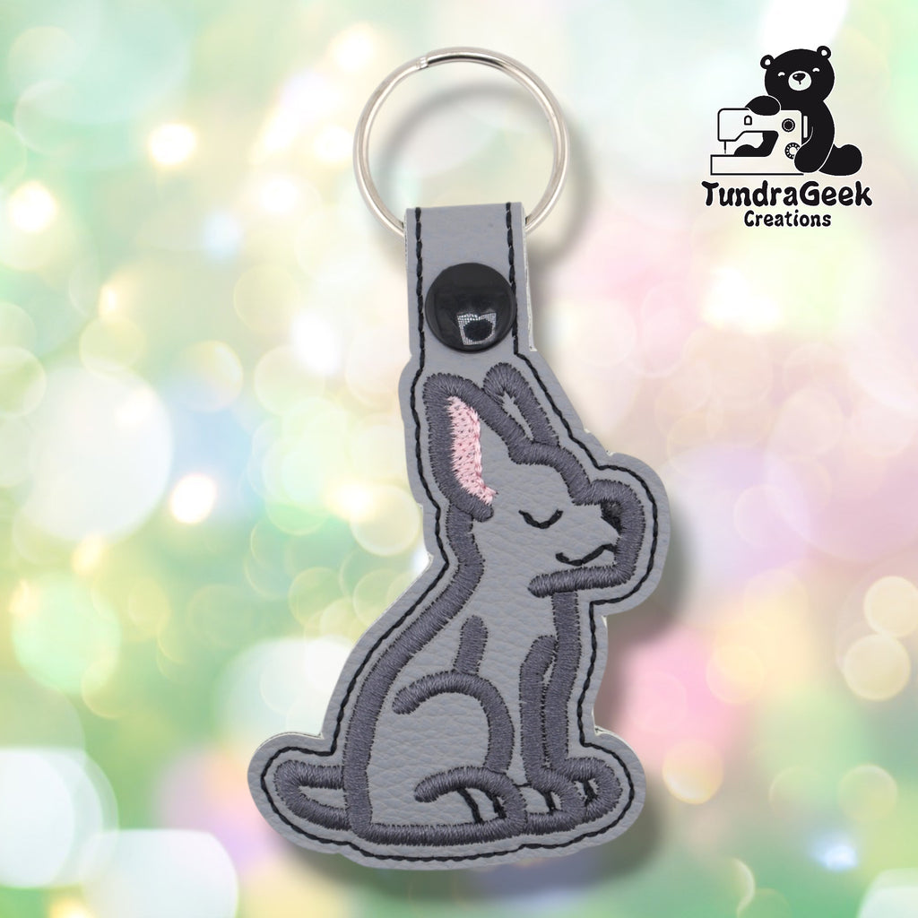Gray embroidered faux leather keychain shaped like a seated Great Dane puppy. Dark gray embroidery details the body and features; pink inner ears and closed eyes are visible. Black snap closure and silver key ring at top. TundraGeek Creations logo in upper right corner.