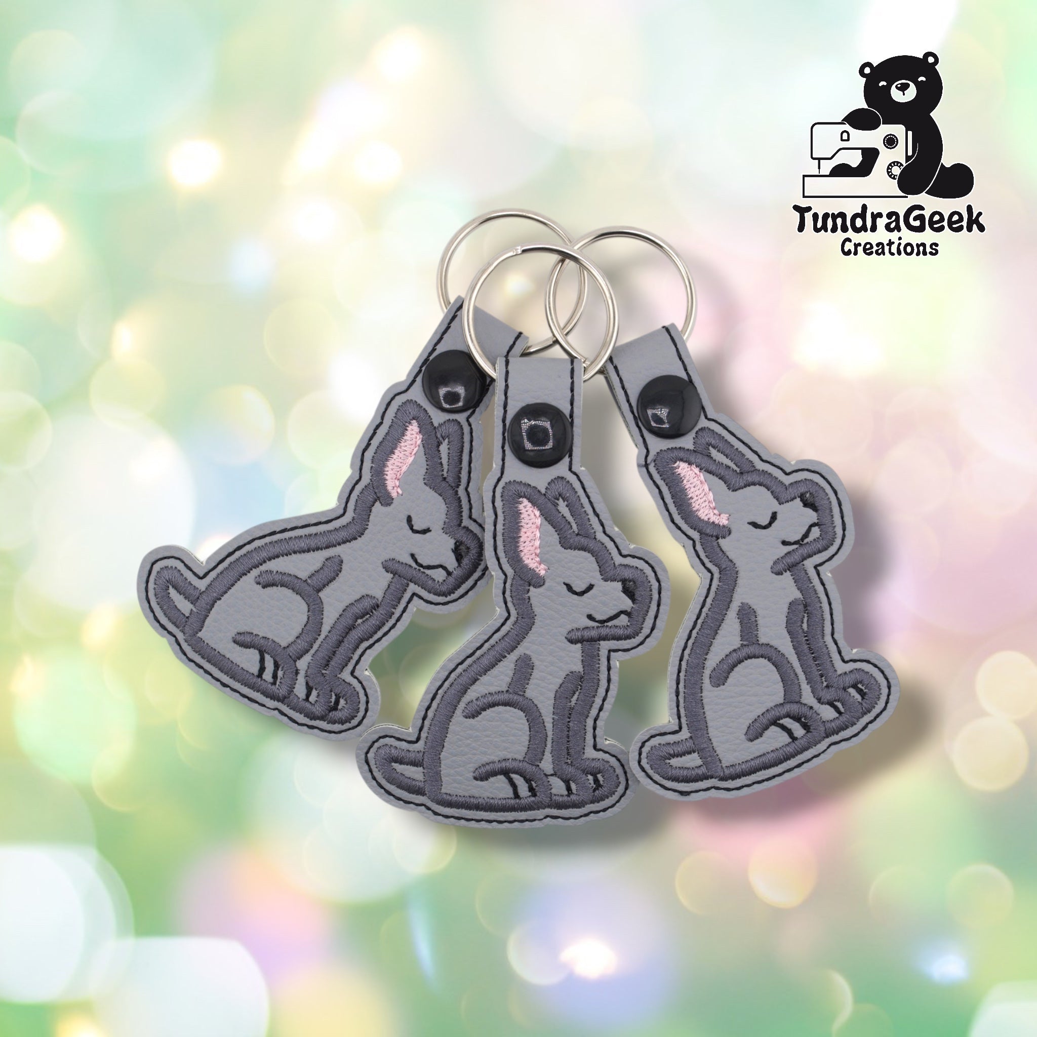 Three Great Dane keychains arranged together on a pastel bokeh background, showing the front face, three-quarter view, and side profile. All three are identical in color and design.