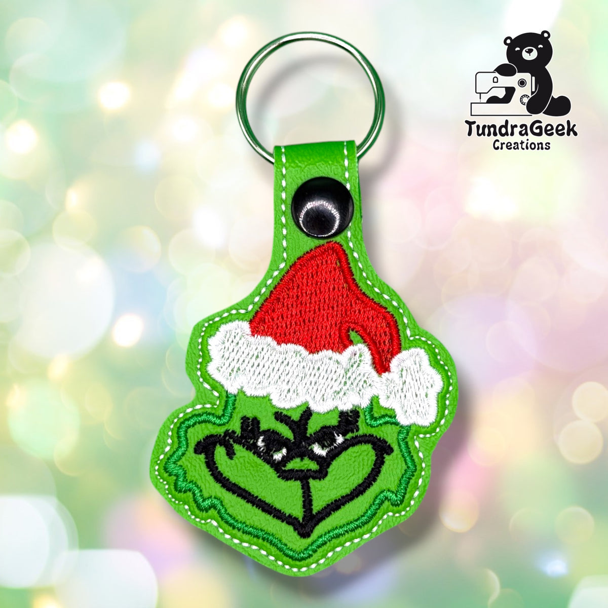 A green Grinch face keychain with a red Santa hat and black outline, displayed against a light bokeh background. Logo in the corner reads TundraGeek Creations with a bear and sewing machine.