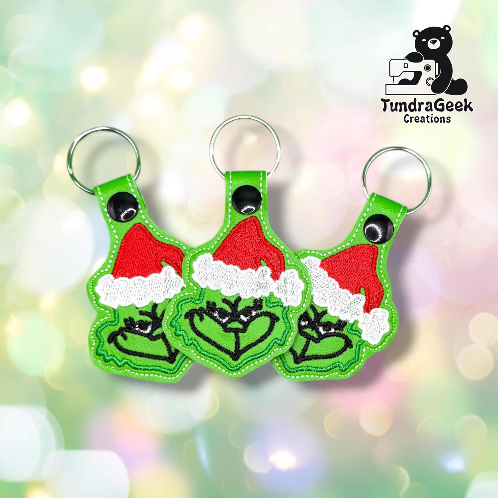 Three green keychains depicting a cartoon character in a Santa hat forming a heart with its hands, displayed against a light bokeh background. TundraGeek Creations logo in the top right corner.