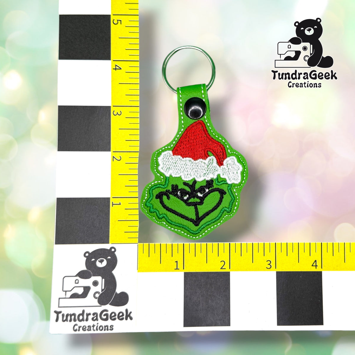 A green keychain with a red Santa hat design is placed next to a ruler for scale, with the TundraGeek Creations logo in the background.