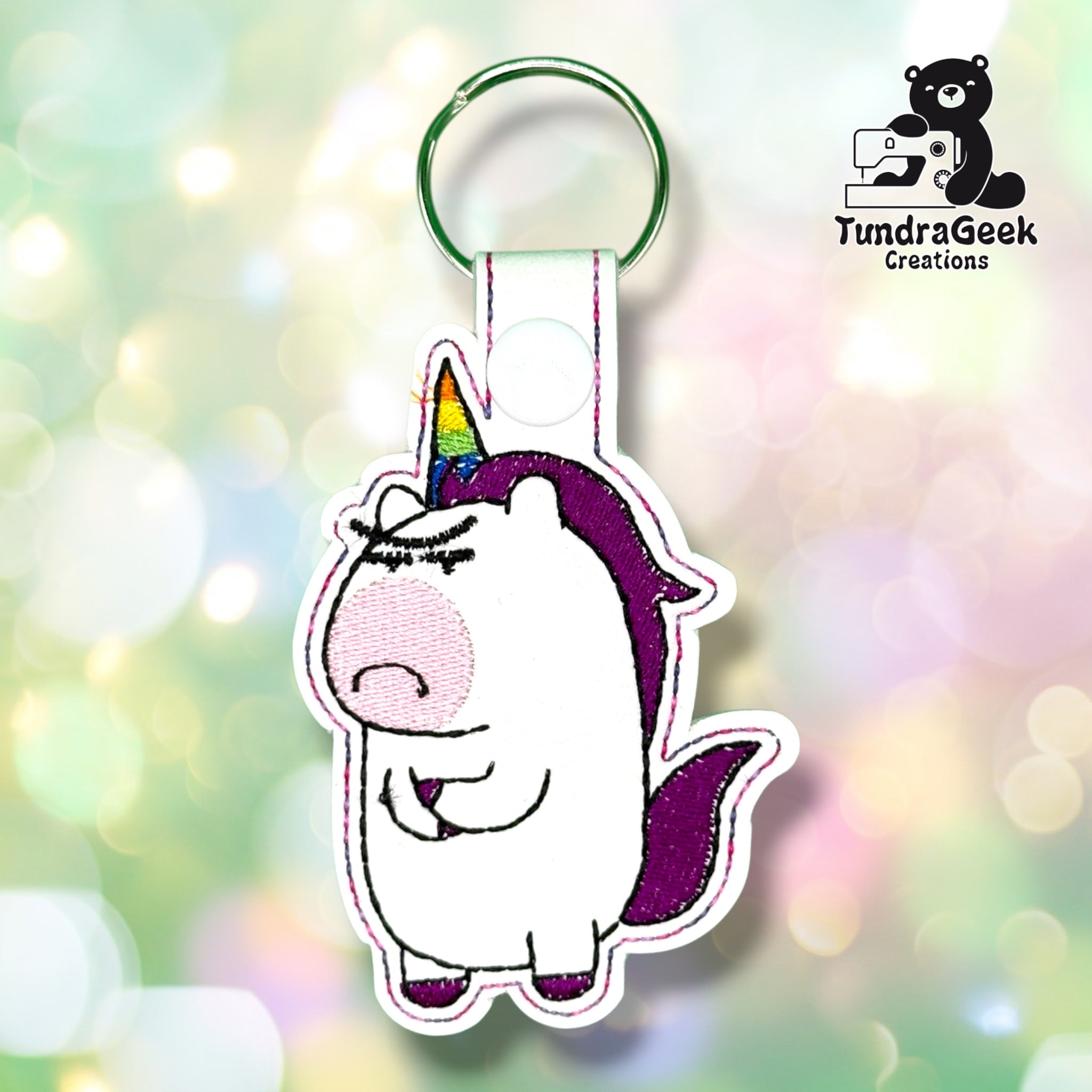 Front view of a grumpy-looking cartoon unicorn embroidered on white vinyl, with a rainbow horn in orange, yellow, green, and blue, a purple mane and tail, purple hooves, a pink cheek, and a heavy-lidded annoyed expression, finished with pink and purple stitched edges and a white plastic snap closure.