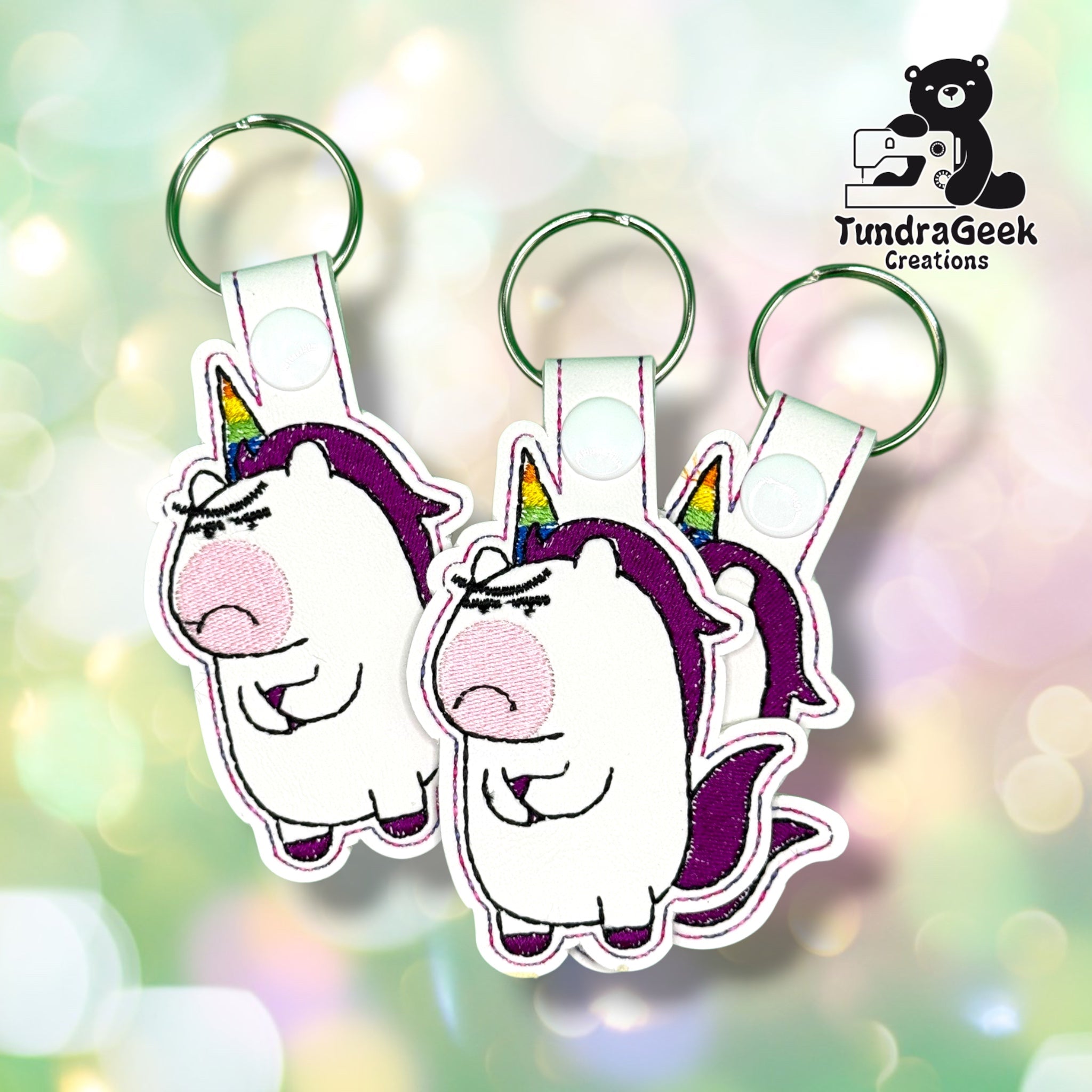 Three of the same keychain shown overlapping together, each with the same grumpy unicorn design in white vinyl with a rainbow horn, purple mane and tail, pink cheek, white plastic snap closures, and green split rings.