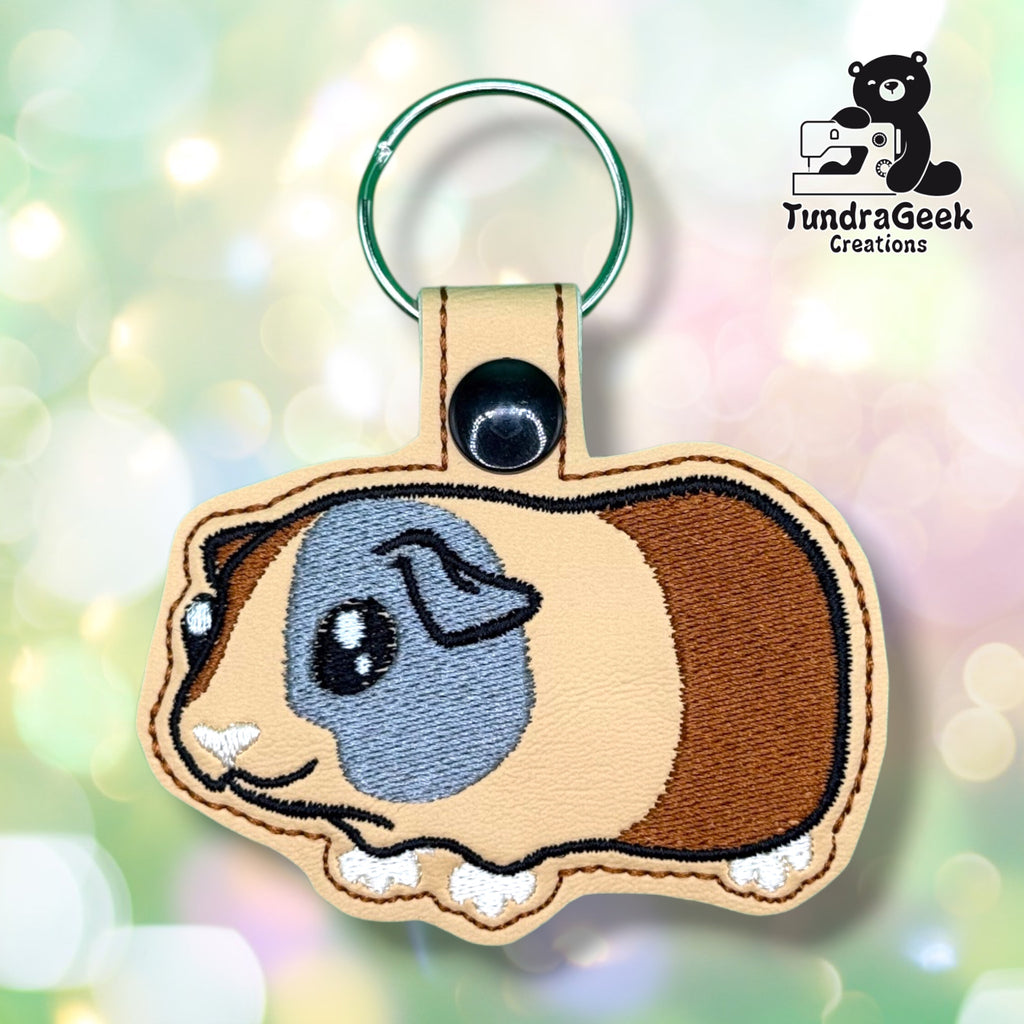 Front of a tan vinyl keychain die-cut in the shape of a guinea pig. Embroidered with a brown patch over one eye and ear, a brown patch running down the body, large soft gray-blue eyes, a pink nose, and small white paws along the bottom edge. Green metal split ring and black snap closure at top.