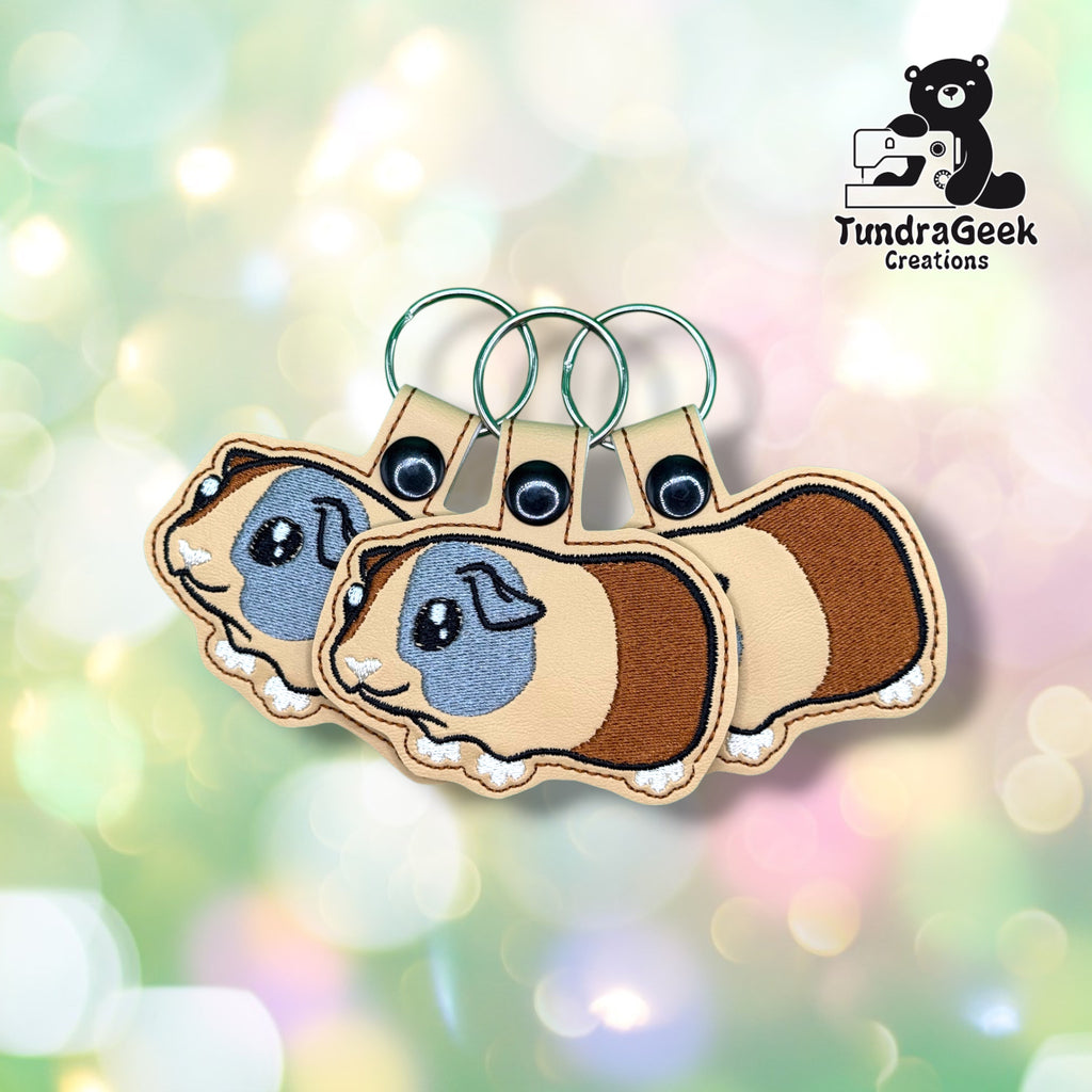 Three identical guinea pig keychains fanned out together, each on green metal split rings, showing the same tan vinyl body with brown patch markings and soft gray-blue eyes.